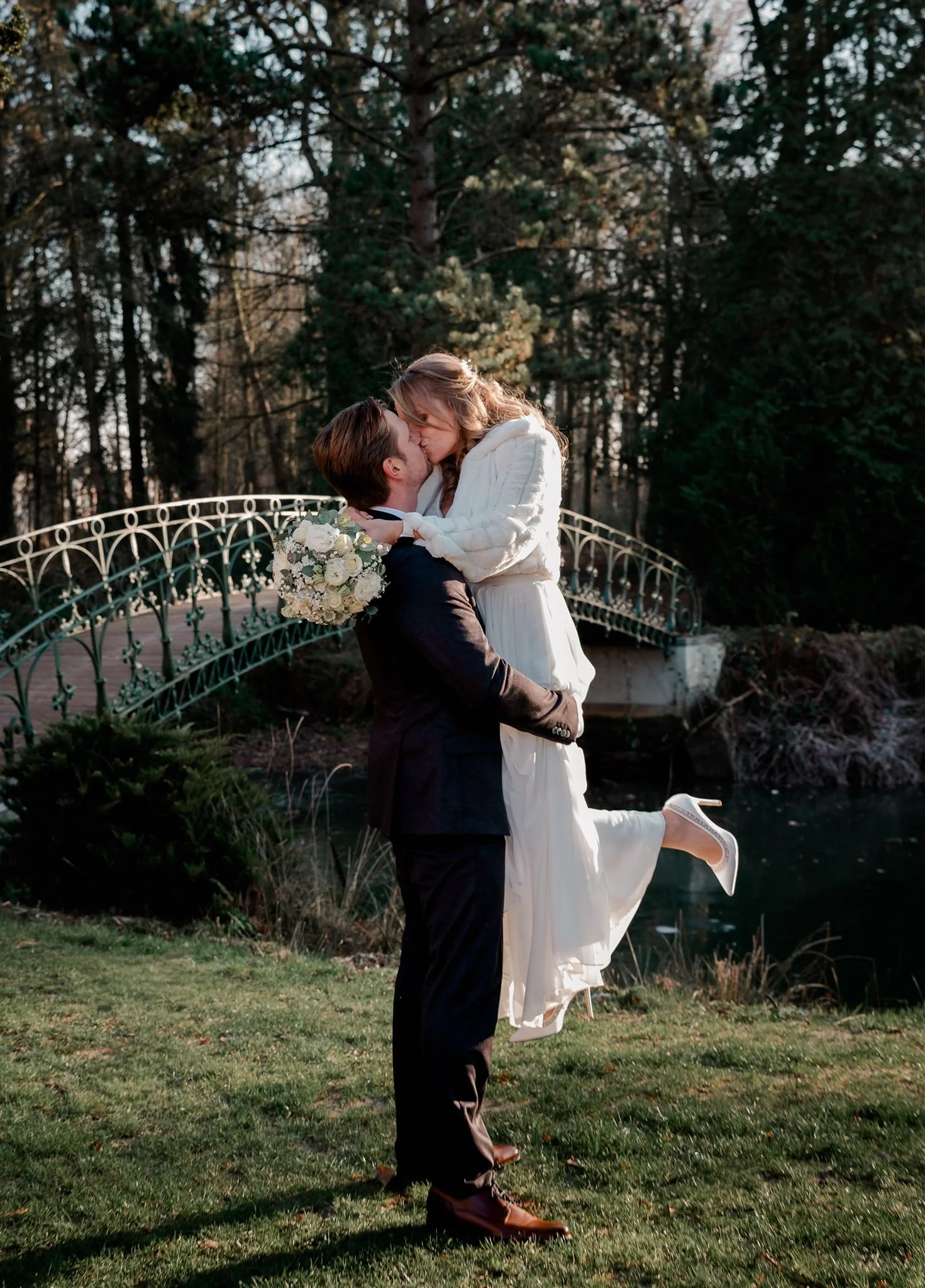 Een stel danst buiten in een park, met een brug en bomen op de achtergrond. Ze lijken te genieten van een romantisch moment.