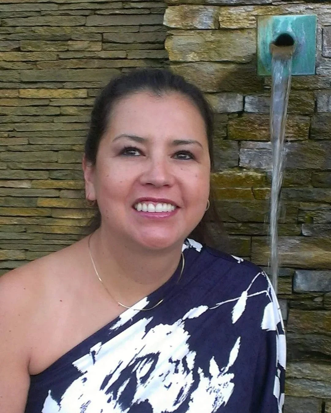 A smiling woman with dark hair, wearing a black and white patterned top, is standing in front of a stone wall with a water feature.