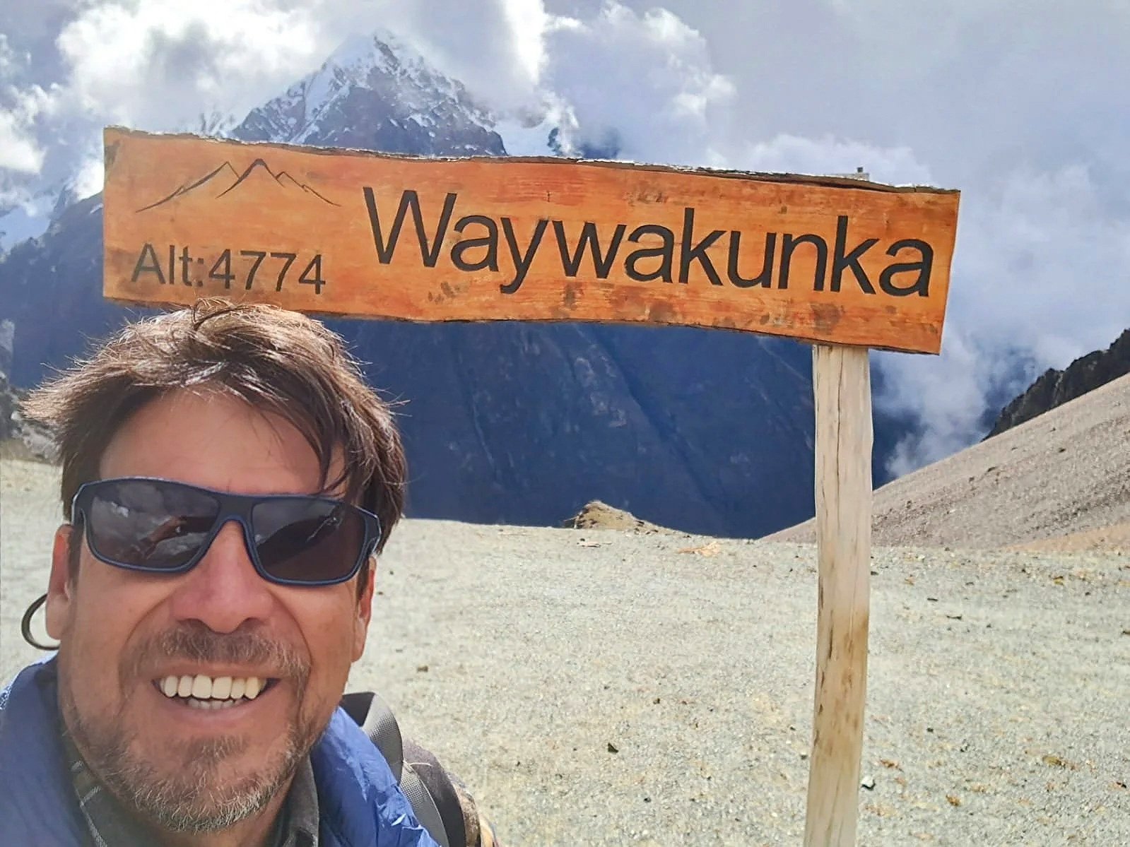 A man wearing sunglasses and a blue jacket taking a selfie near a wooden sign that reads 'Waywaku nka' at an altitude of 4774 meters, with snow-capped mountains in the background.