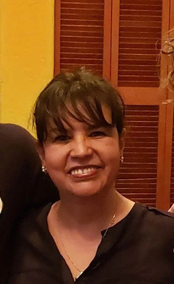 Smiling woman with short dark hair, wearing earrings and a black top, in front of louvered wooden doors and a yellow wall.