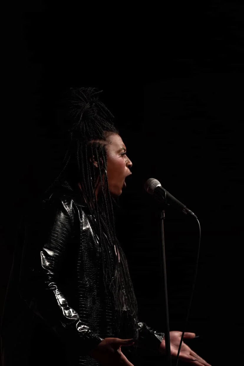 A woman with braided hair speaking passionately into a microphone against a black background.