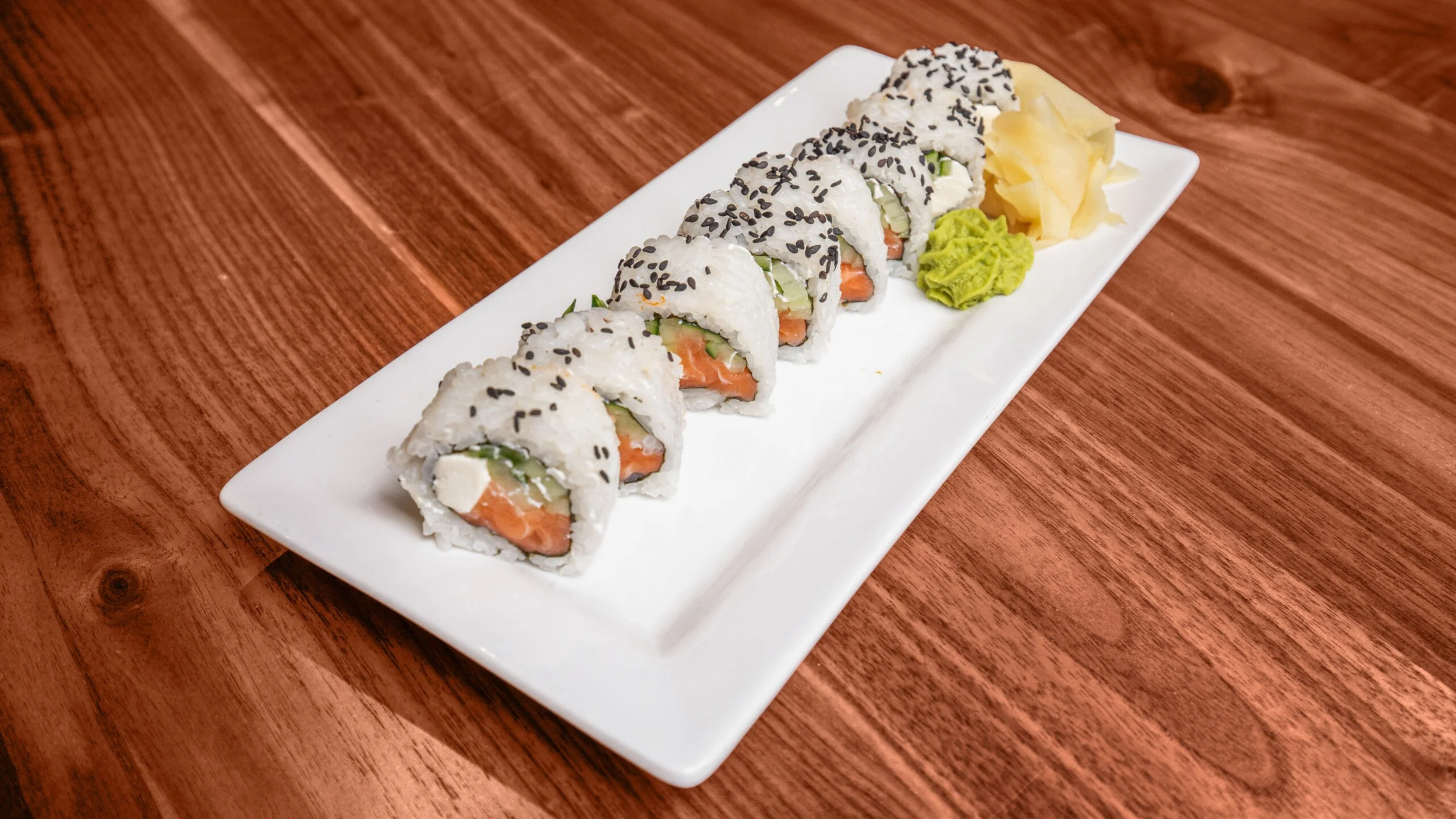 A rectangular white plate with seven sushi rolls, garnished with black sesame seeds, accompanied by wasabi and pickled ginger on a wooden table.