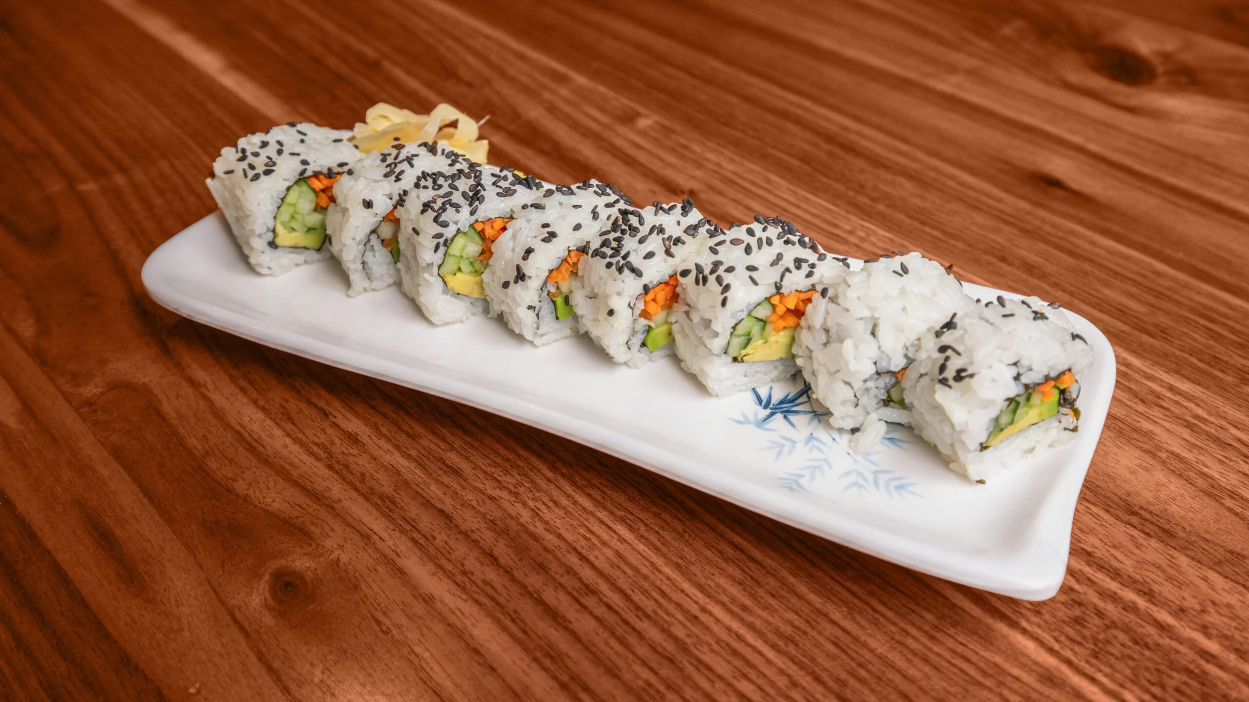 Sushi roll slices with cucumber, carrot, and possibly avocado, topped with black sesame seeds, arranged on a white rectangular plate on a wooden table.