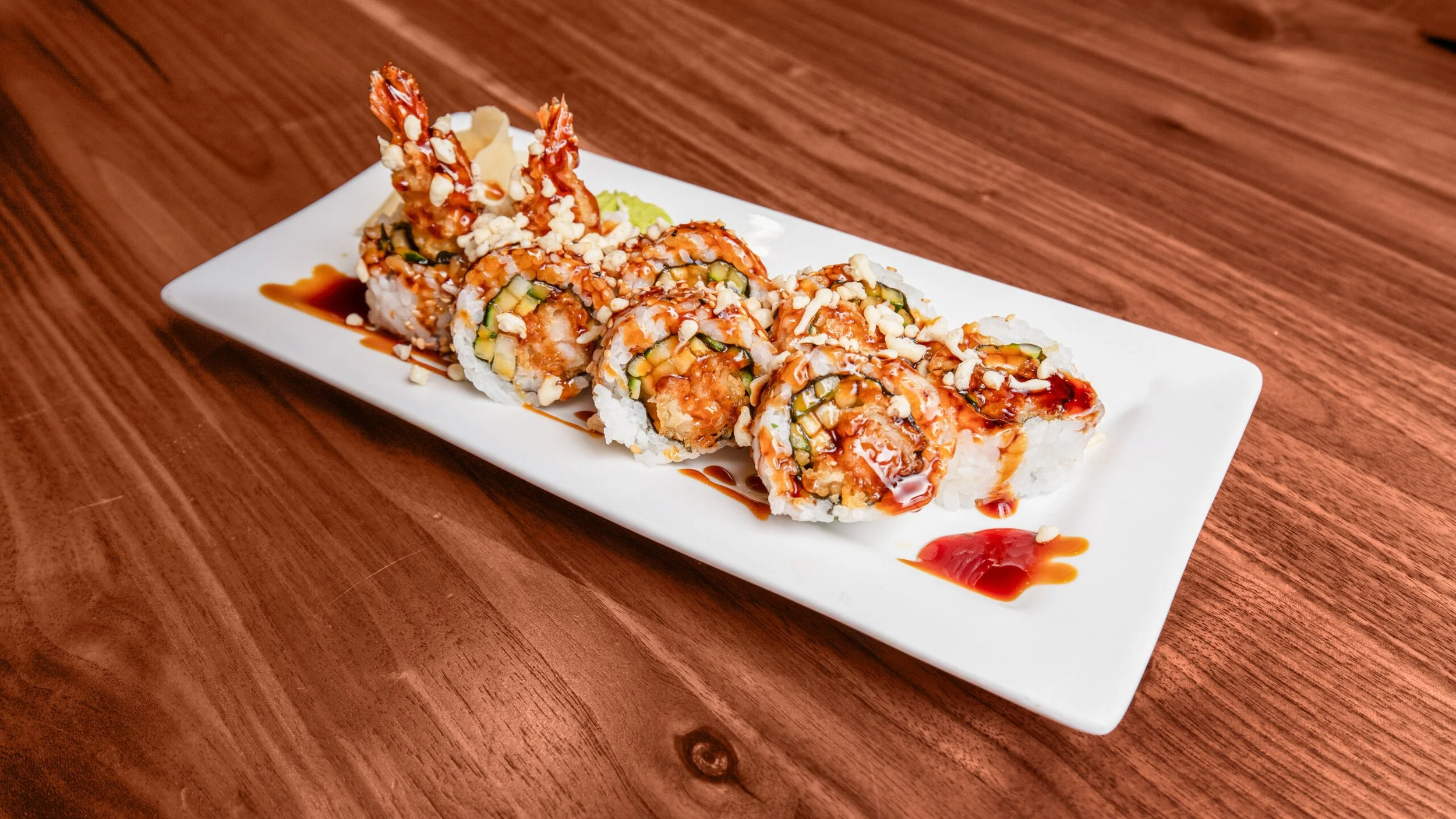 Sushi roll on a rectangular white plate with eel sauce drizzled on top, placed on a wooden table.