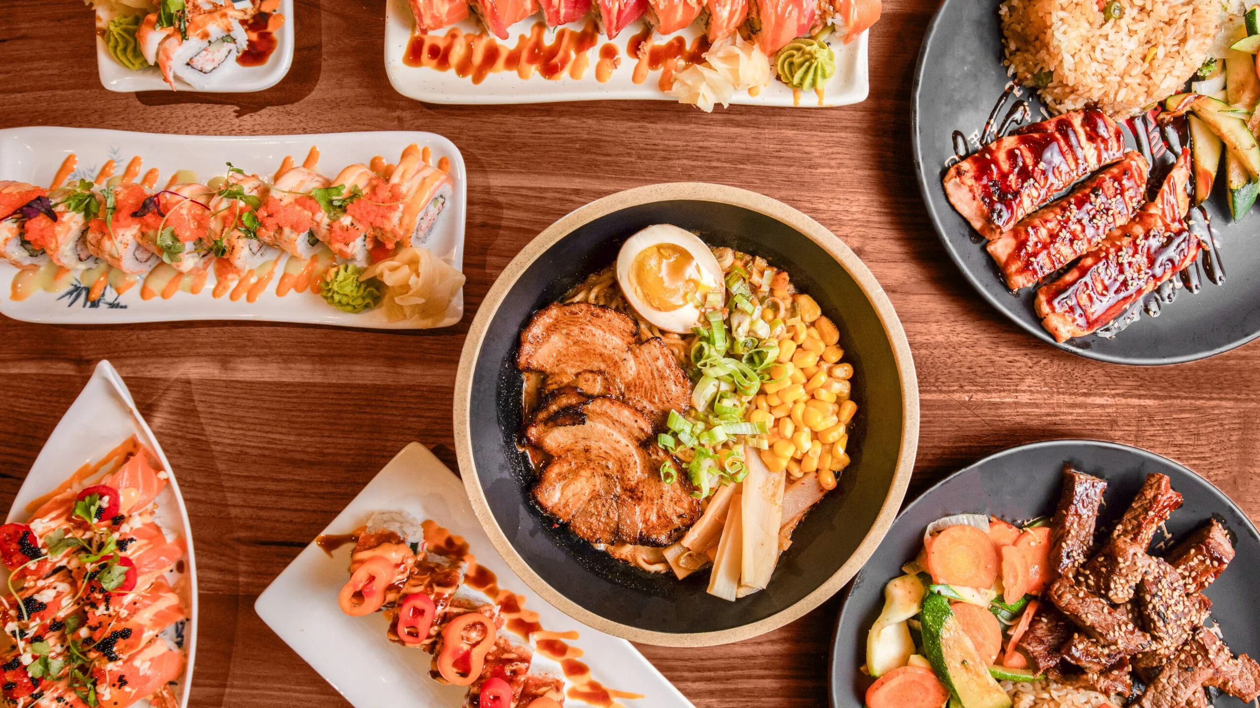 Assorted Japanese dishes on a wooden table, including sushi rolls, ramen bowl with sliced pork, egg, and vegetables, glazed eel, grilled beef, and rice with vegetables.