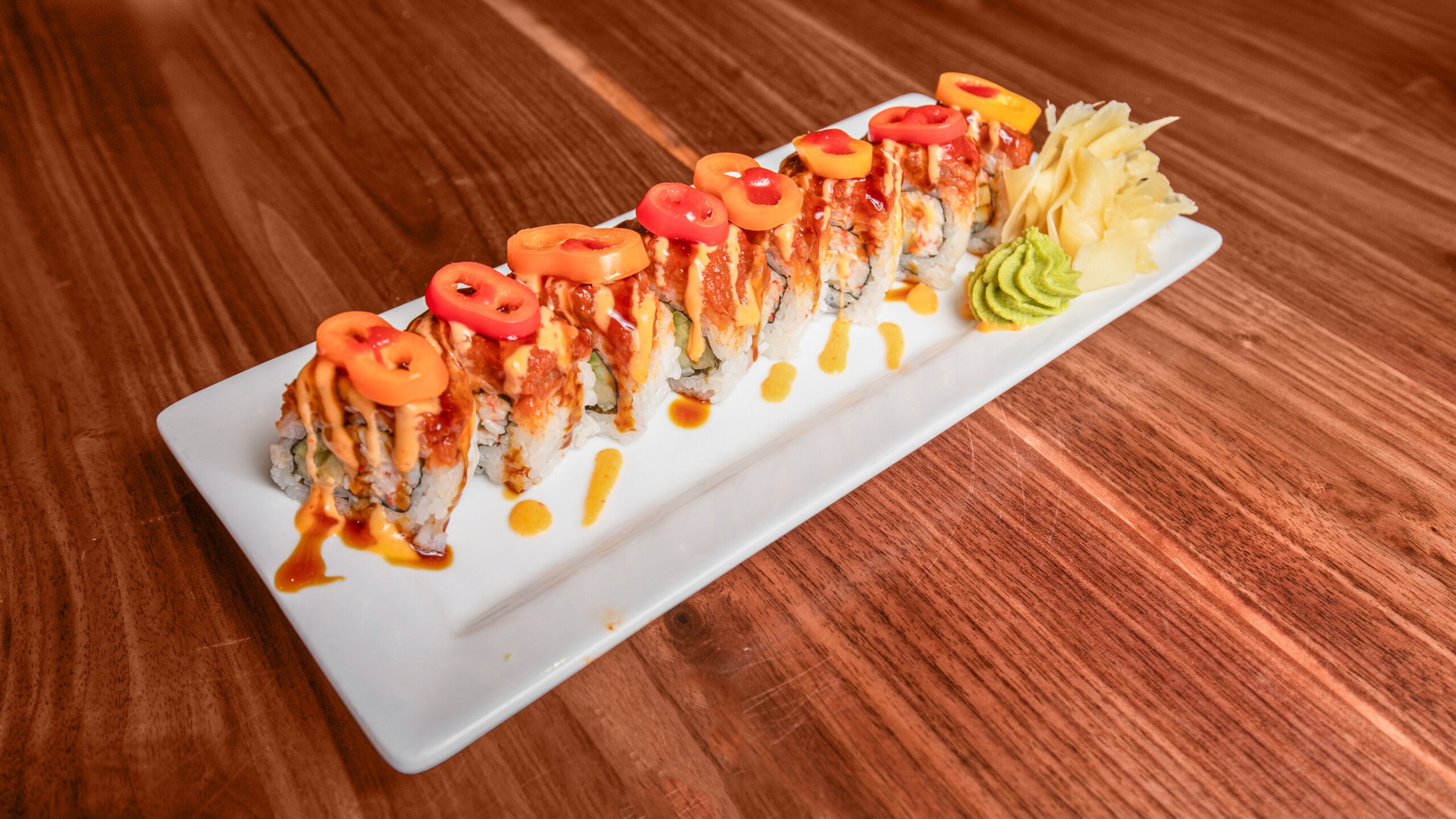 A plate of sushi rolls drizzled with sauces, topped with sliced red and yellow peppers, served with wasabi and ginger on a rectangular white plate, on a wooden table.