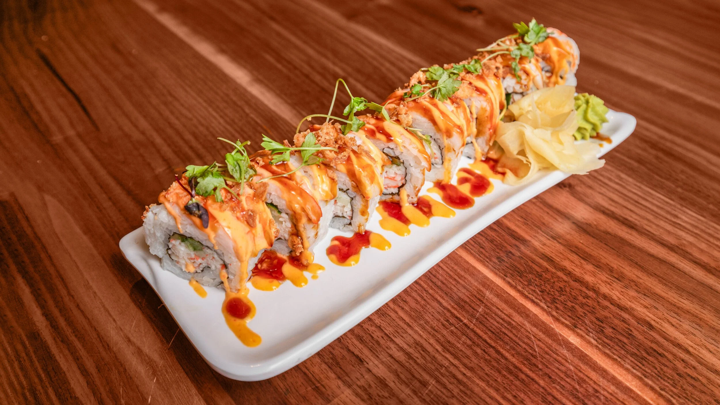 A plate of sushi rolls drizzled with orange and red sauces, garnished with green herbs and accompanied by pickled ginger and wasabi on a wooden table.