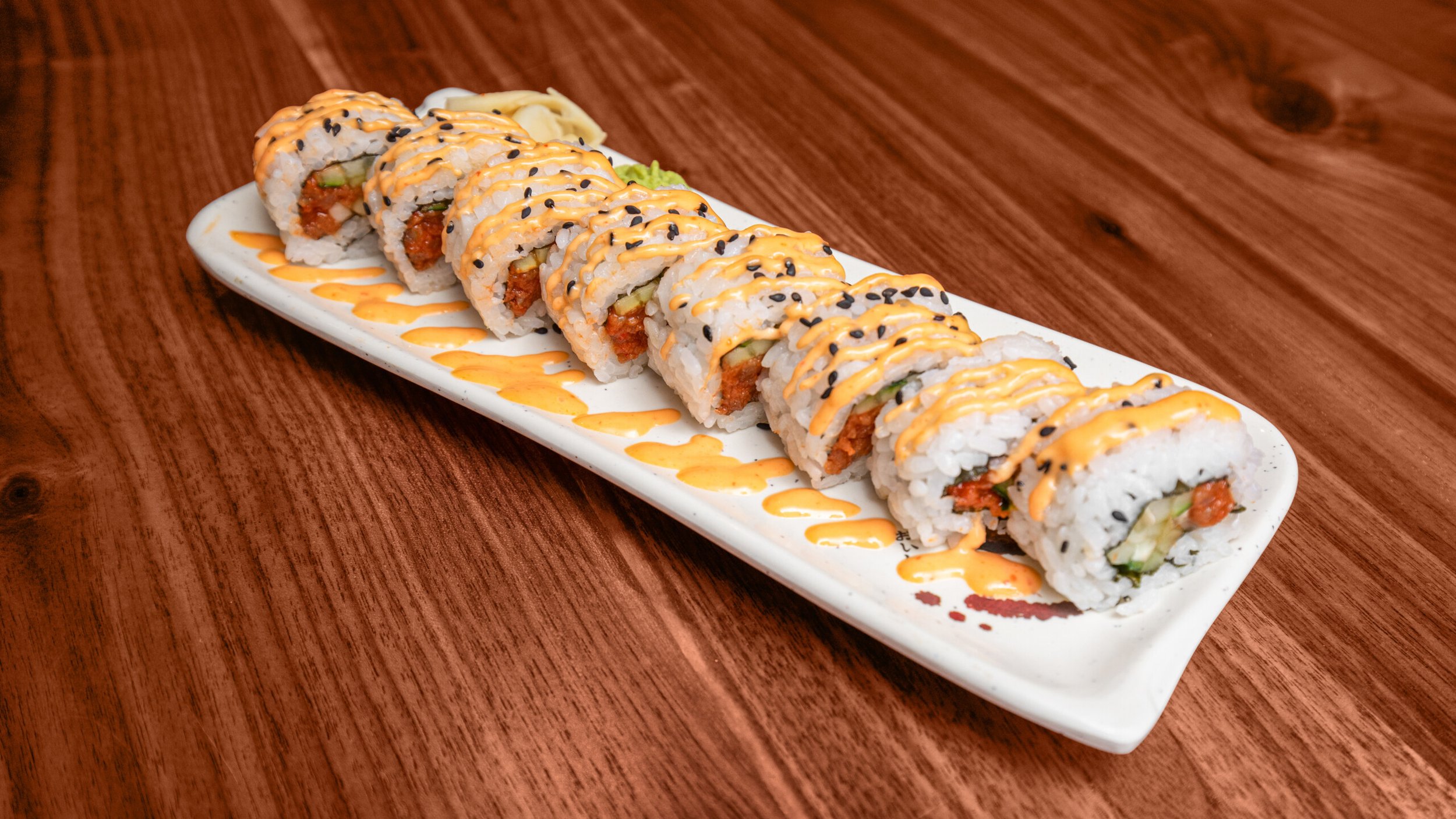 A plate of sushi rolls topped with orange sauce and black sesame seeds, decorated with a drizzle of additional orange sauce, placed on a wooden table.
