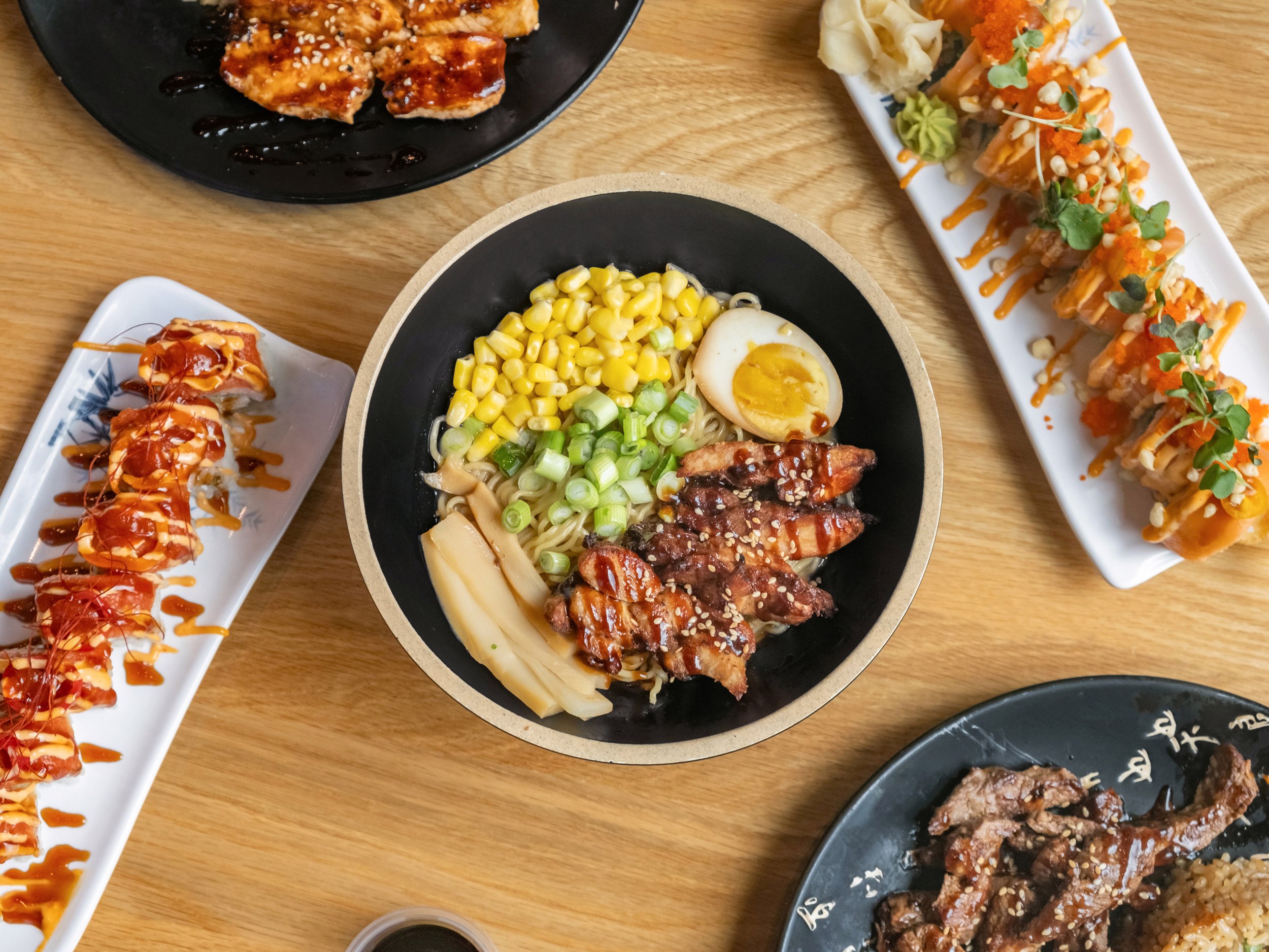 Assorted Japanese dishes on a wooden table, including sushi rolls drizzled with sauce, a bowl of ramen with corn, green onions, bamboo shoots, half a boiled egg, and grilled meat.
