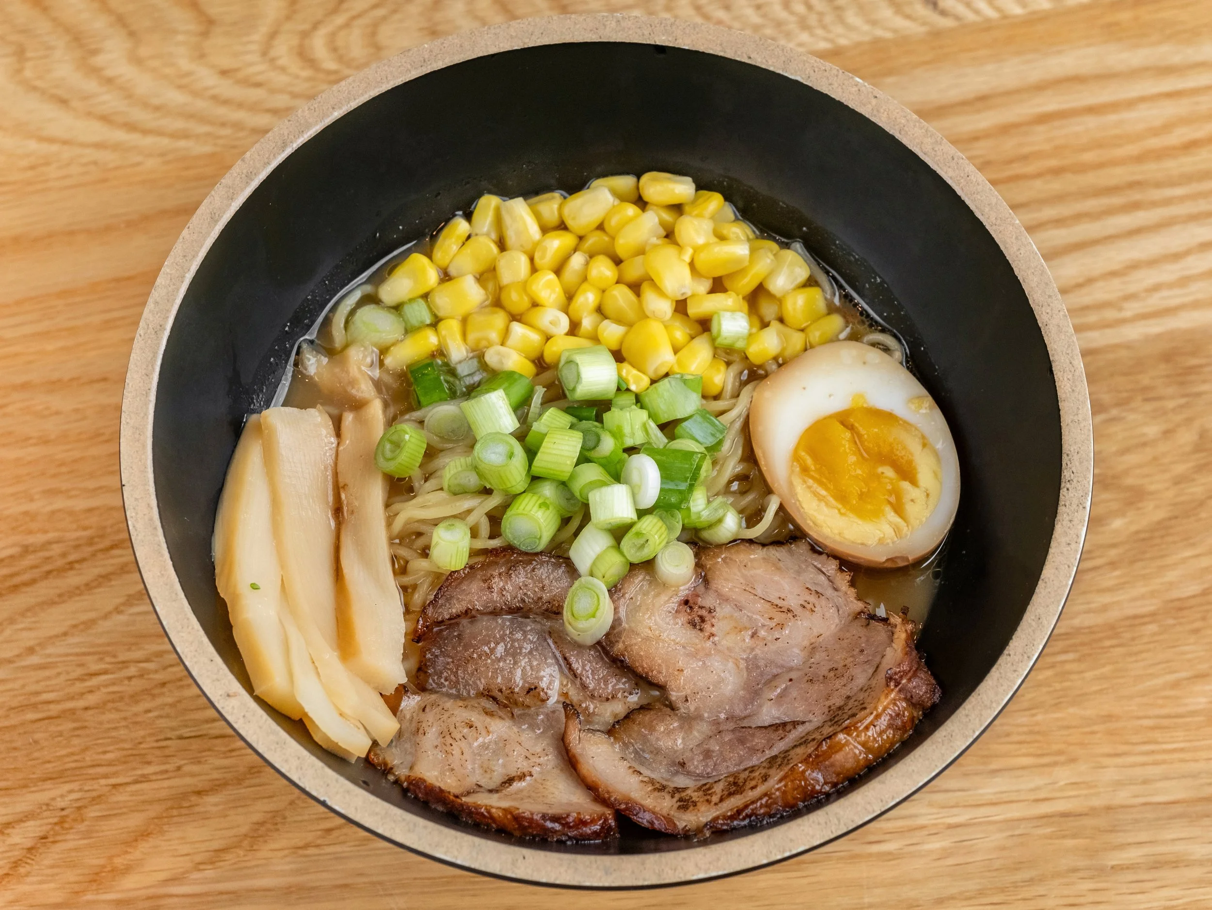 Bowl of ramen with corn, green onions, bamboo shoots, sliced pork, soft-boiled egg, and noodles on a wooden surface.