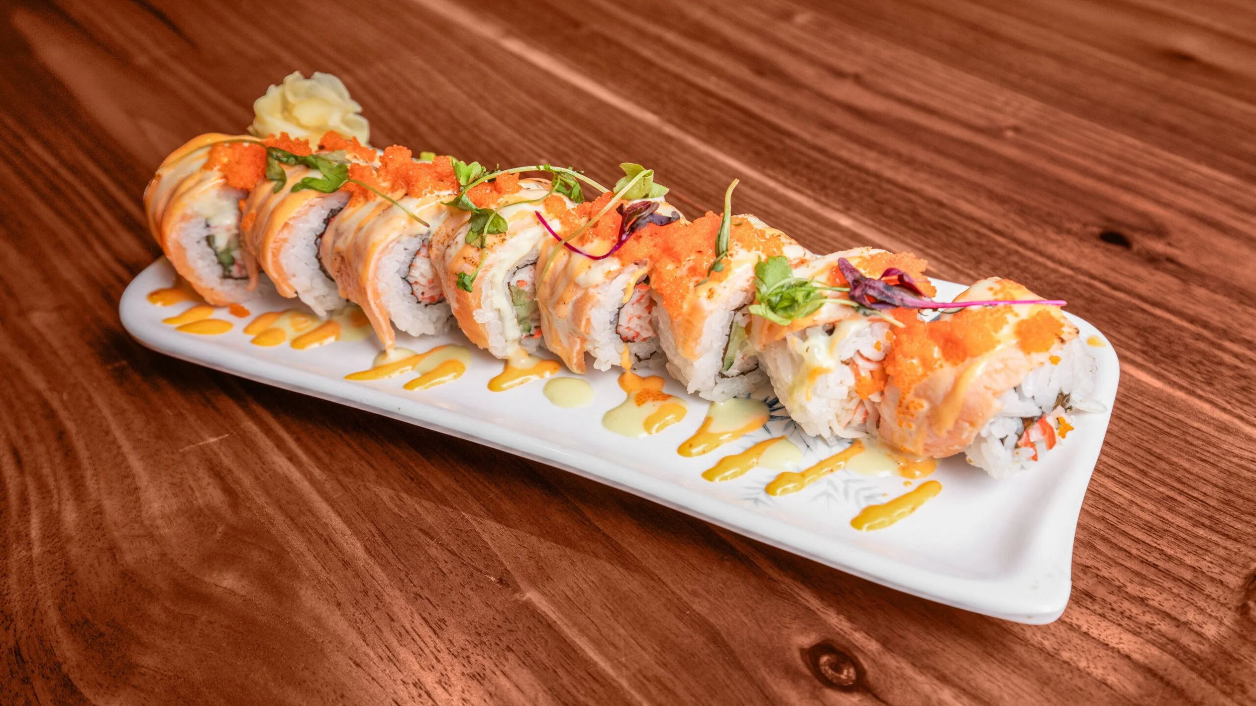 Sushi roll with orange and yellow sauces, garnished with microgreens, on a white rectangular plate on a wooden table.