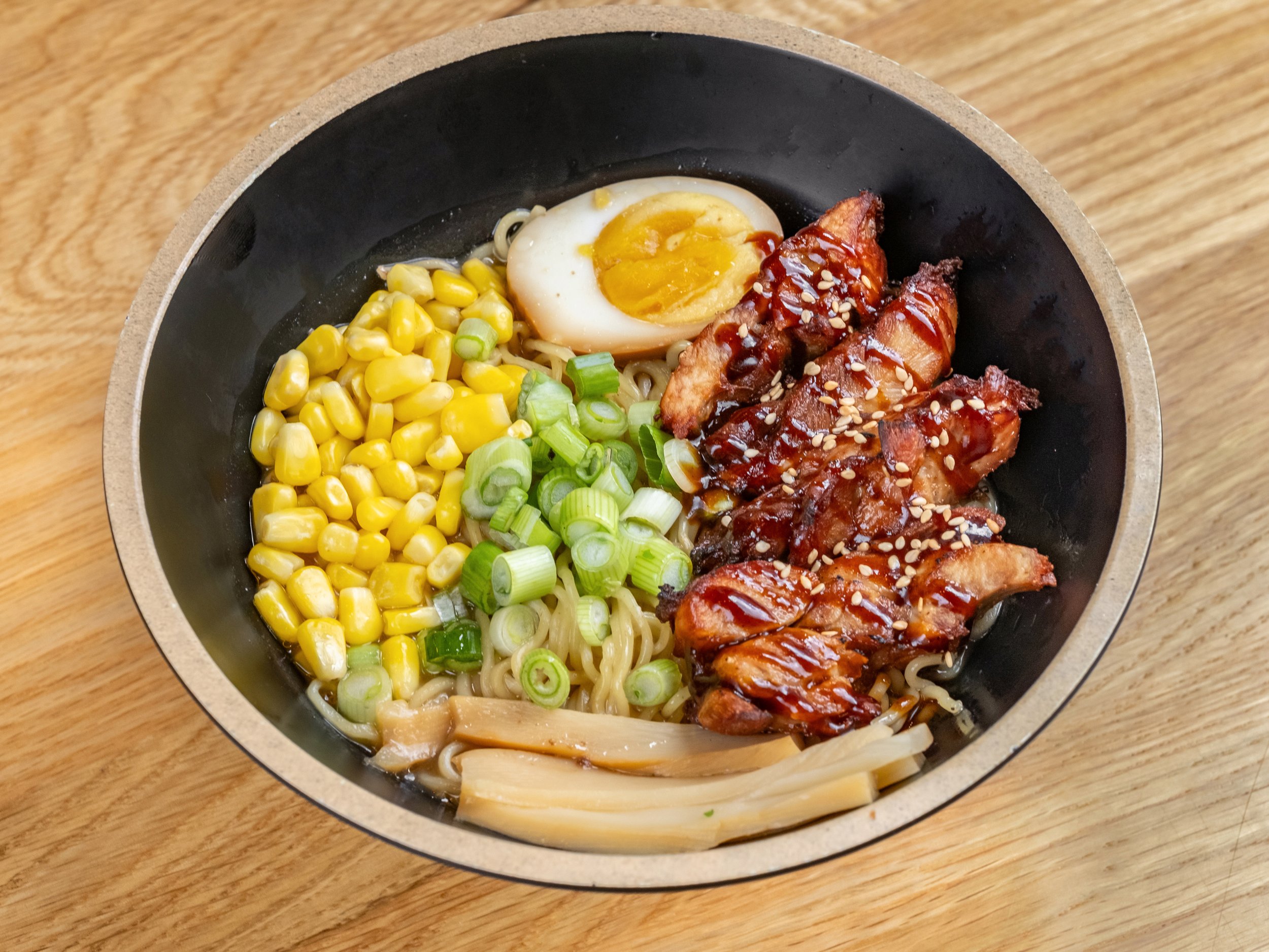 Bowl of ramen noodles topped with sliced green onions, corn kernels, a halved soft-boiled egg, and glazed grilled chicken with sesame seeds.