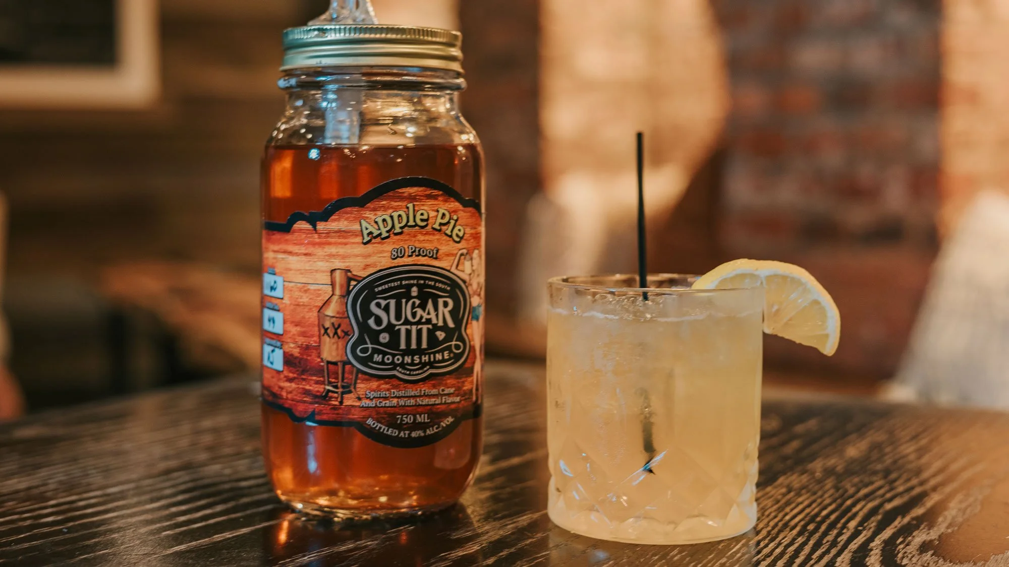 A bottle of Apple Pie flavored moonshine next to a glass of lemonade with ice and a lemon wedge on a wooden table.