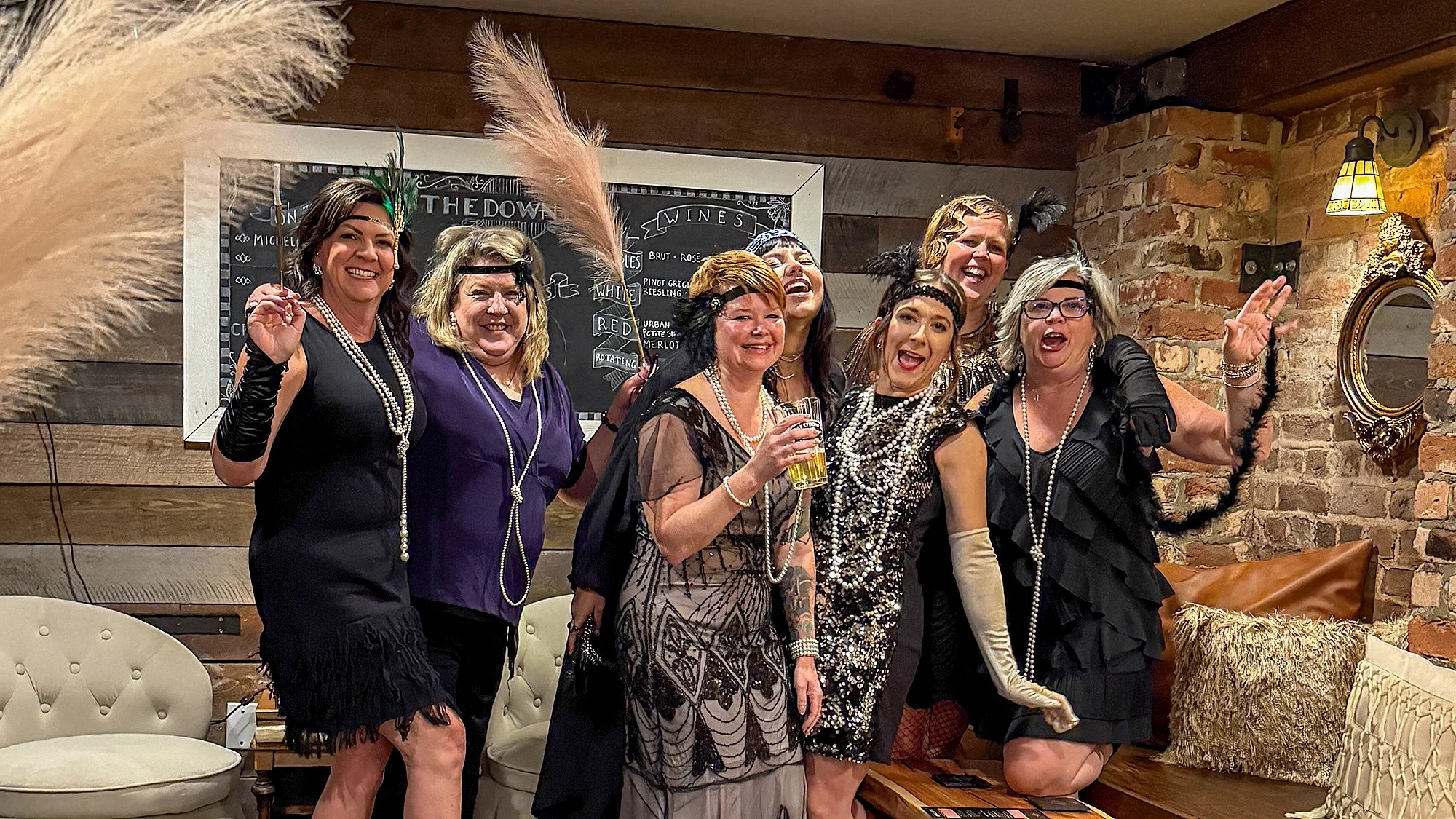Group of women dressed in 1920s-inspired costumes celebrating together in a cozy, rustic setting, some wearing pearl necklaces, feather headbands, and vintage-style dresses, with a brick wall, a chalkboard menu, and warm lighting in the background.