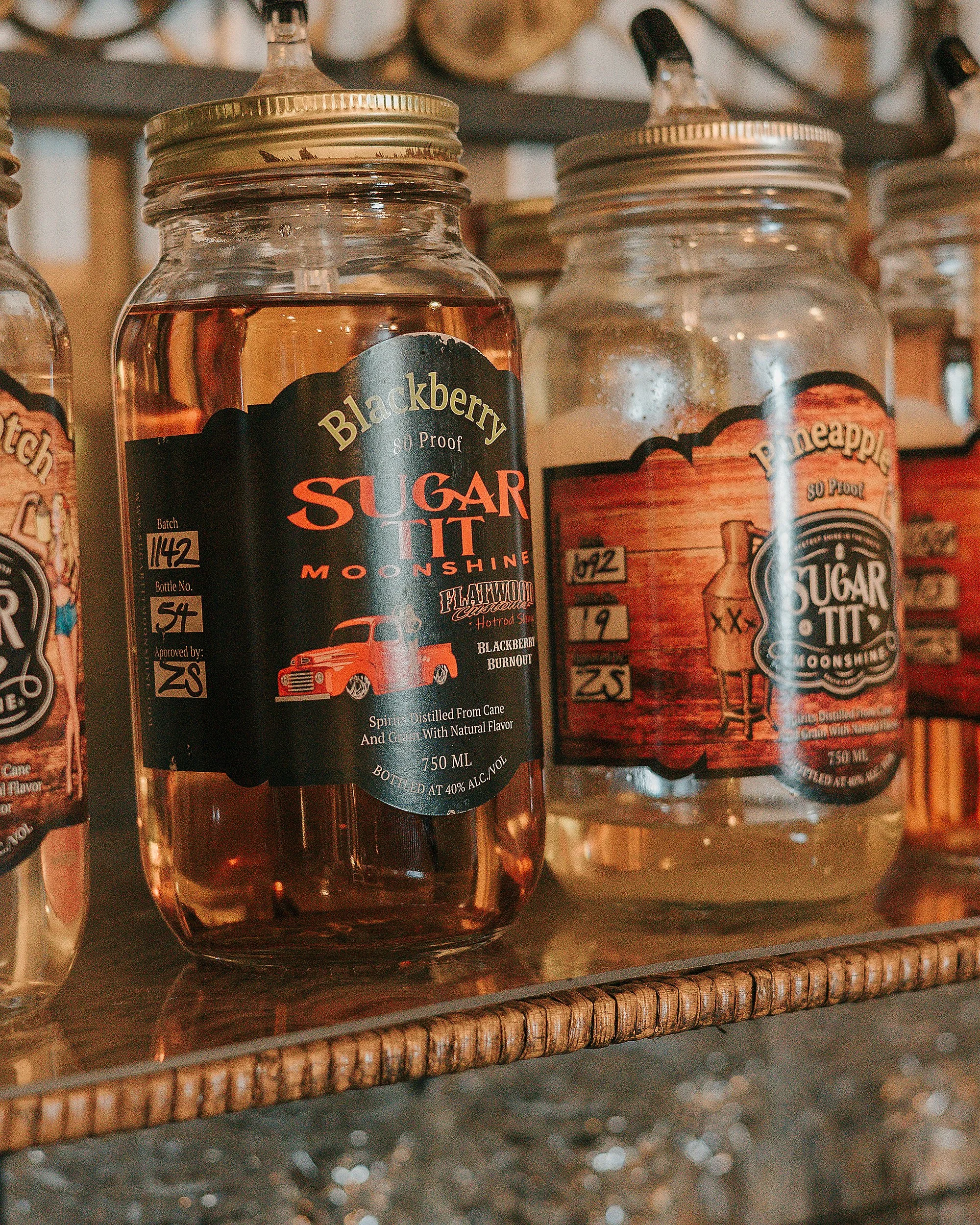 Glass jars on a wooden shelf containing flavored sugars such as blackberry, pineapple, and others, labeled with black and colorful labels.