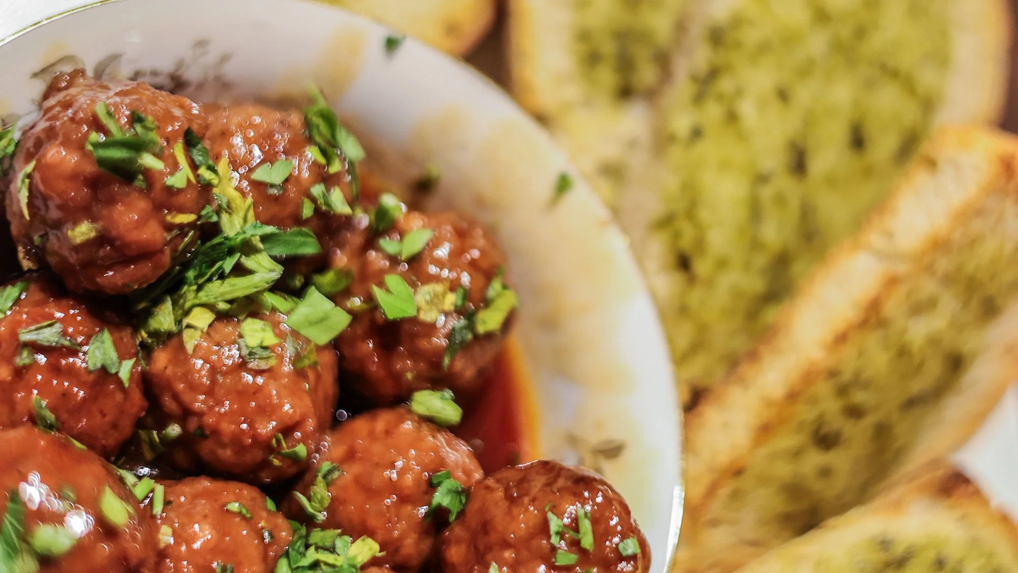 Close-up of glazed meatballs garnished with chopped green herbs on a white plate with grilled bread in the background.