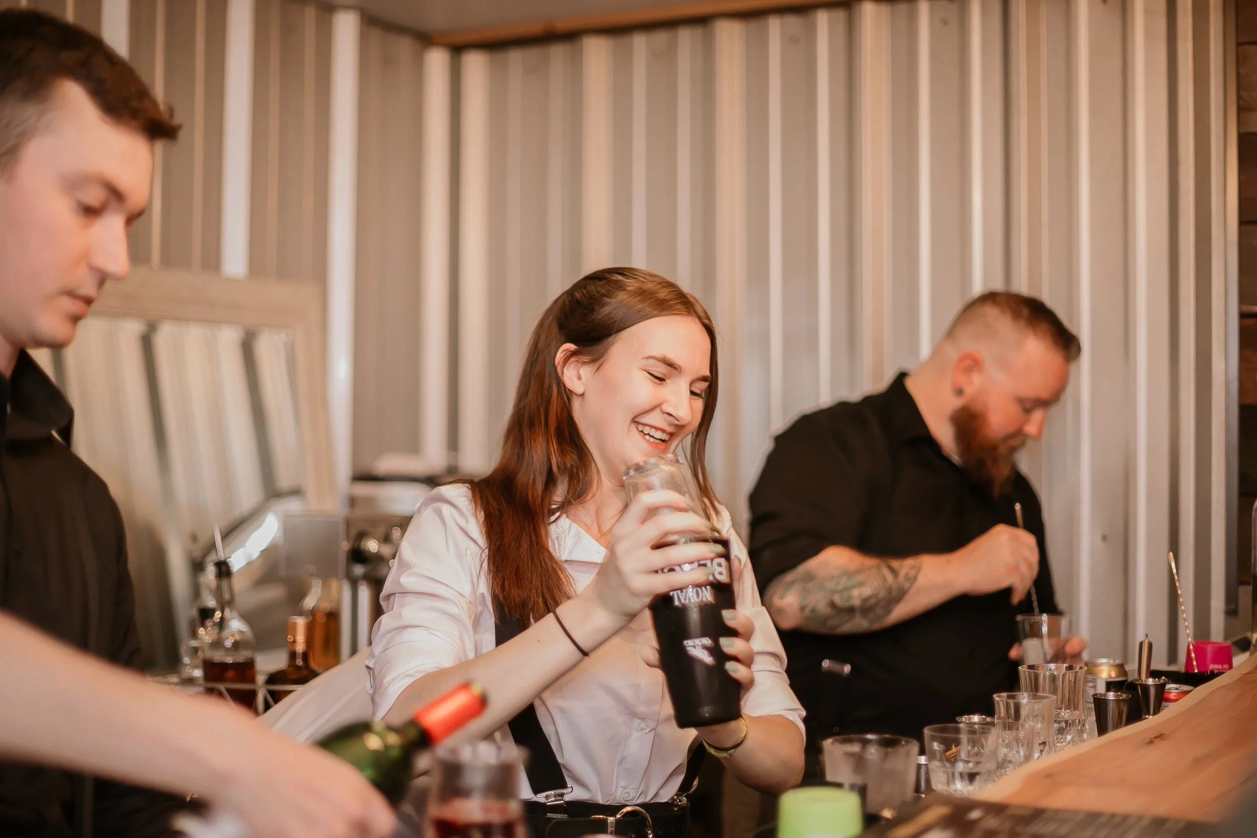 A woman with long reddish-brown hair smiling and holding a dark beverage in a clear glass jar, sitting at a bar or counter with two men on either side, in a setting with vertical striped wall panels.