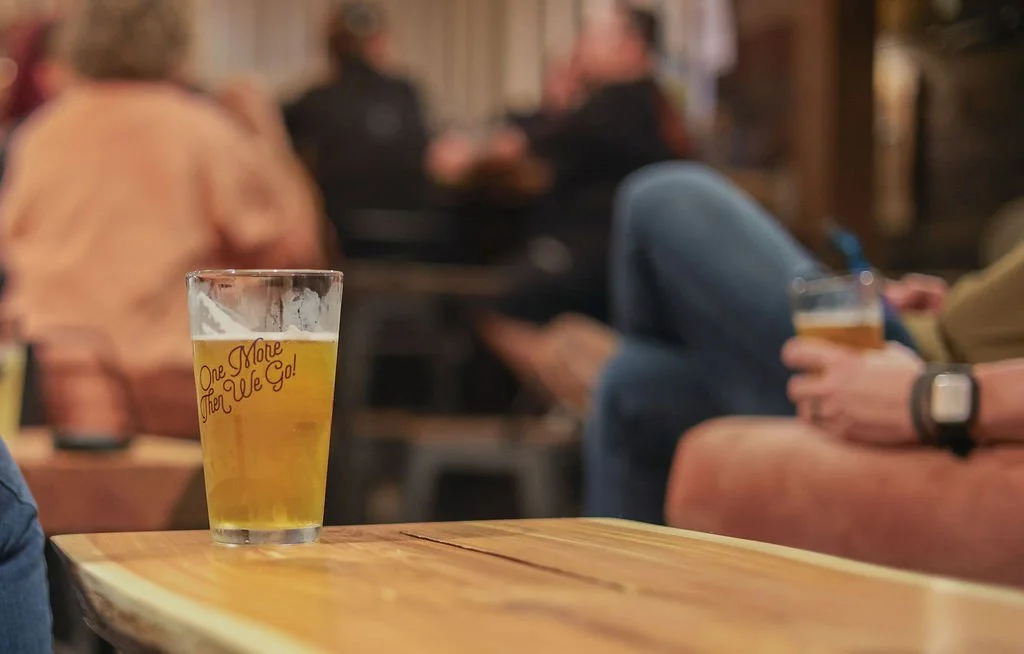 A glass of beer on a wooden table with the phrase "One More Then We Go!" written on it, while people in the background sit and chat in a casual setting.