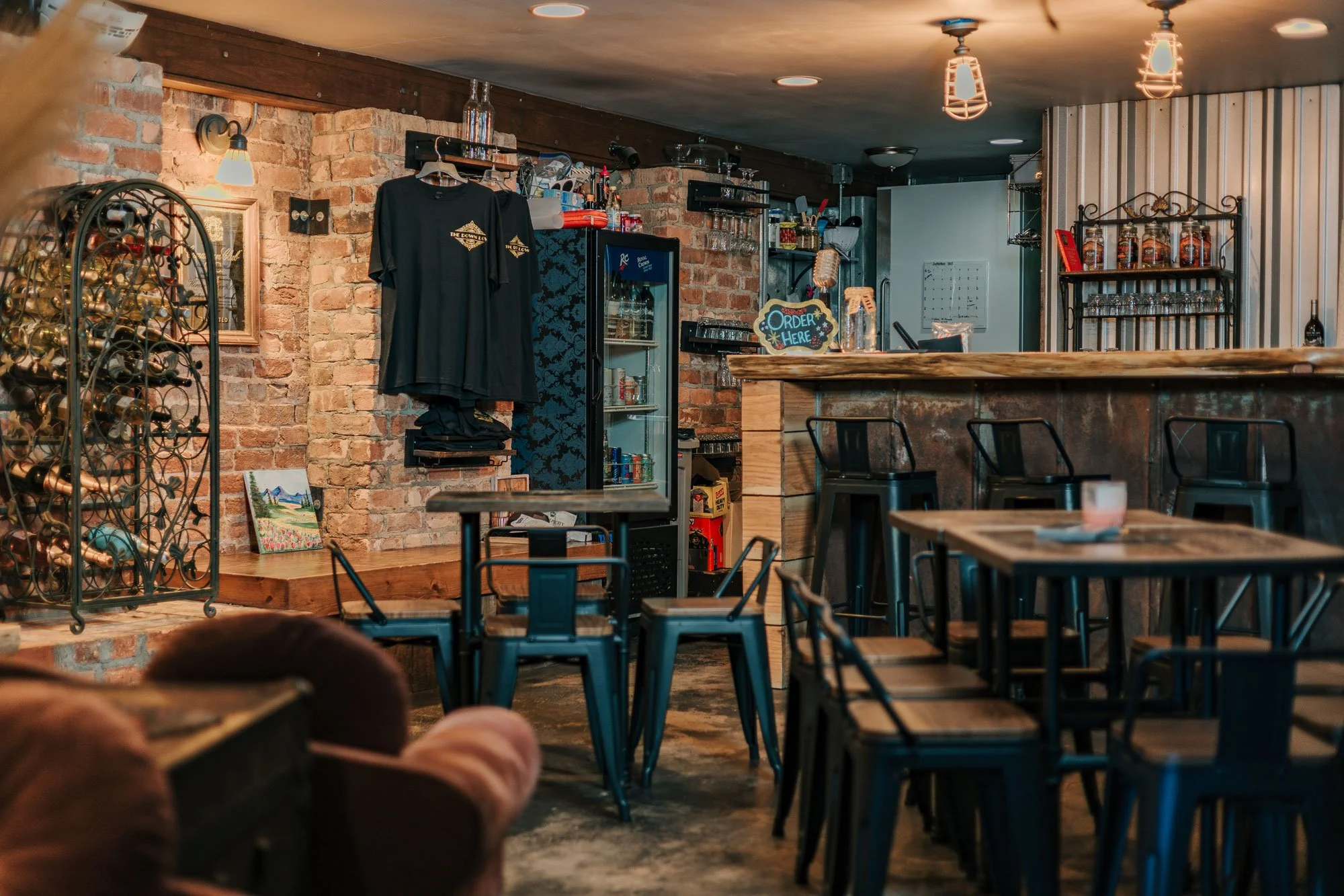 Interior of a cozy restaurant or bar with wooden tables and black chairs. The decor includes exposed brick walls, vintage lighting fixtures, and shelves with bottles and glasses. There is a sign that says 'Order Here' near the bar area. Part of a person's feet can be seen in the foreground.
