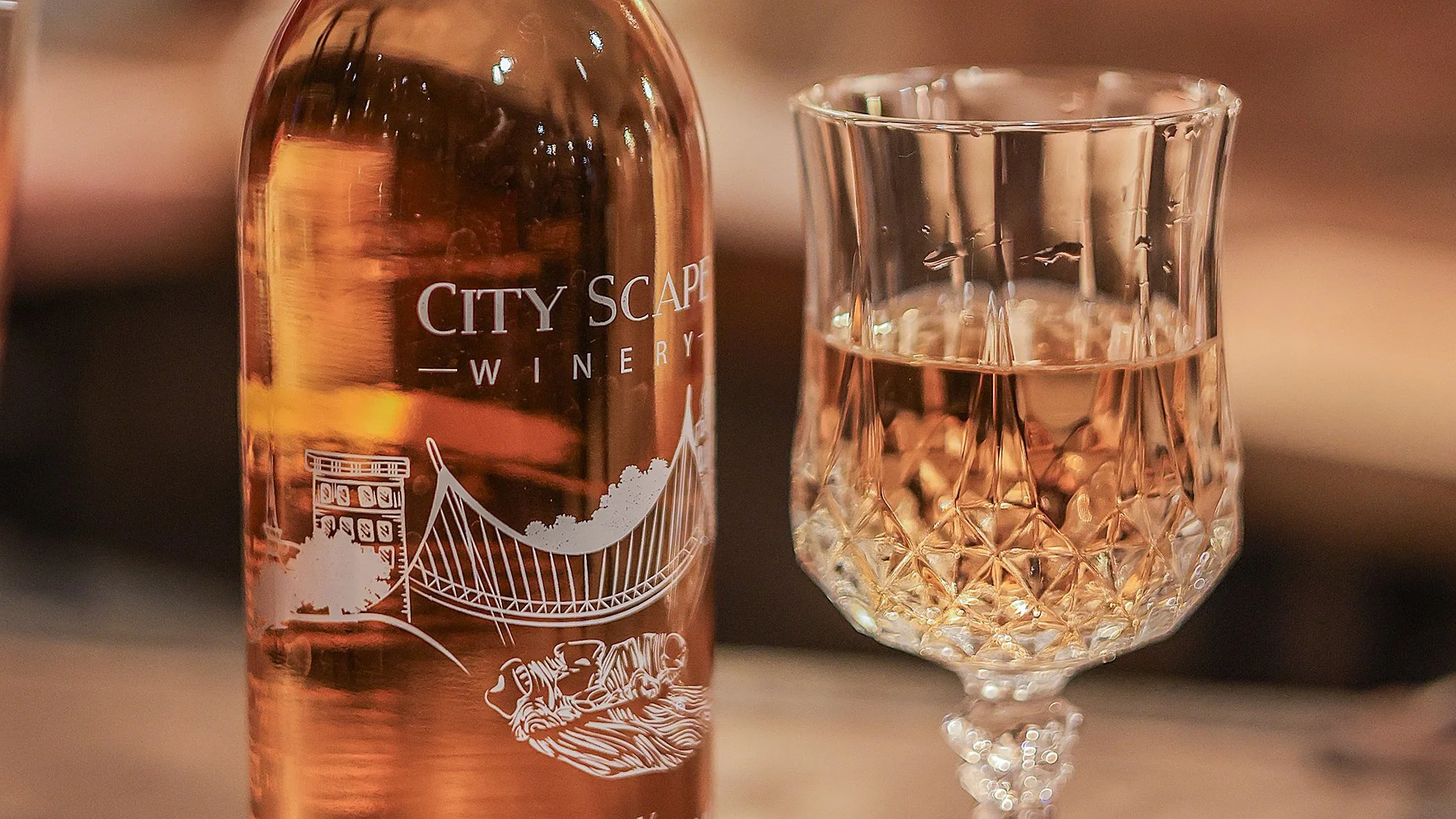 A partially visible pink bottle labeled 'City Scape Winery' next to a clear glass with a diamond pattern filled with a light pink liquid.
