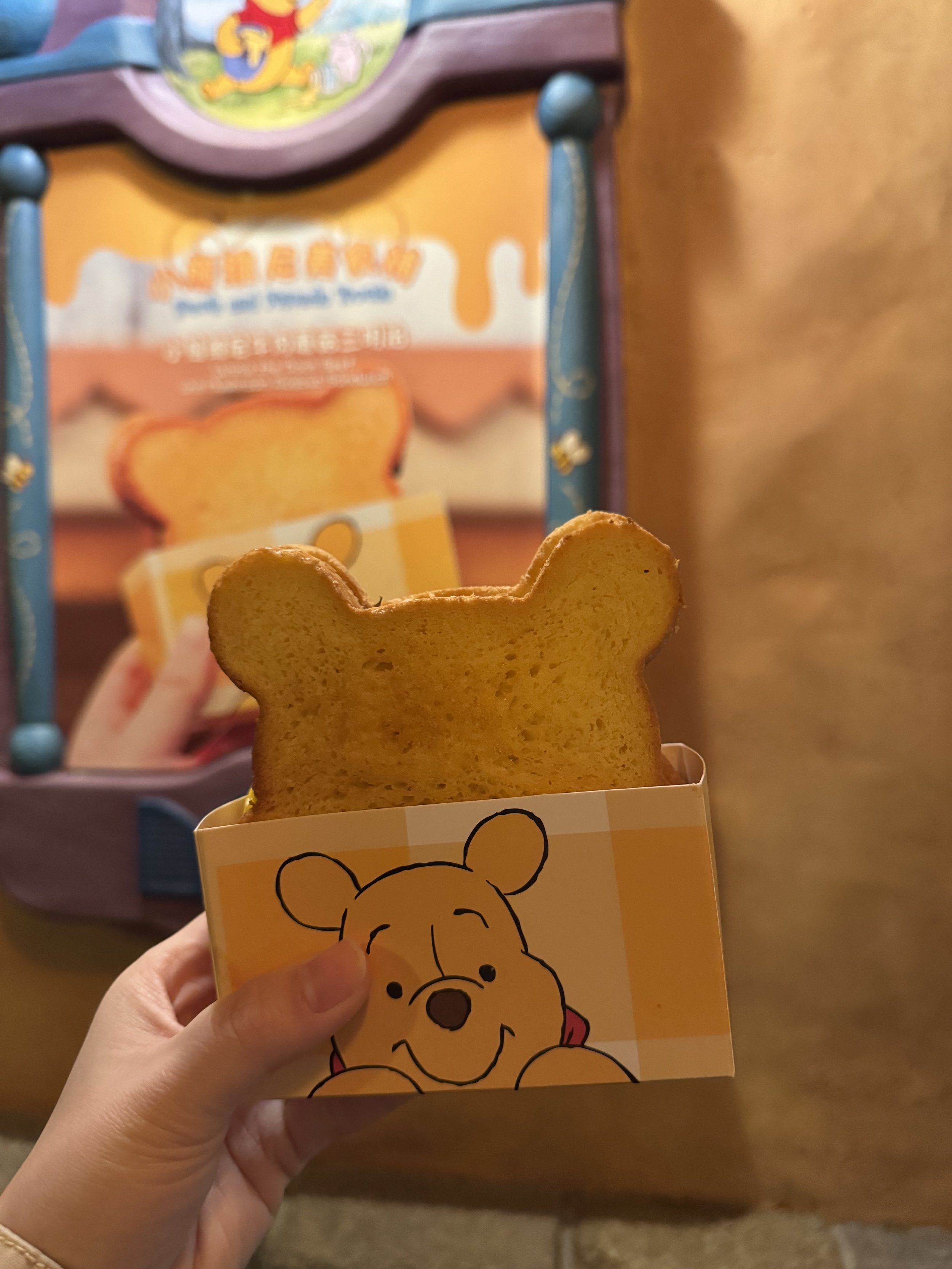 Winnie the Pooh shaped sandwich in a Winnie the Pooh paper sleeve in front of a building with a poster image of the sandwich