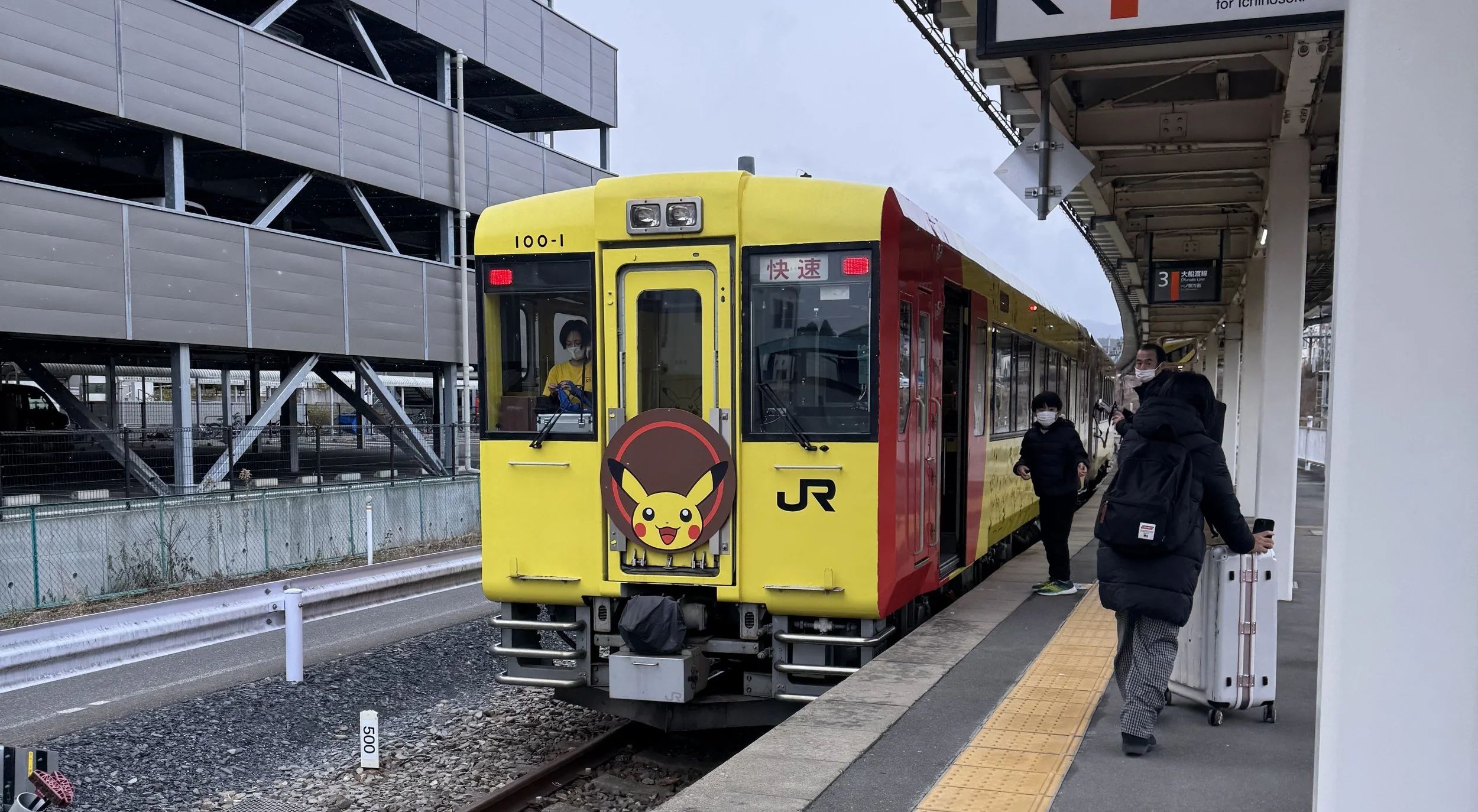 Yellow JR train with a Pikachu on a circular sign in the front stopped at a station