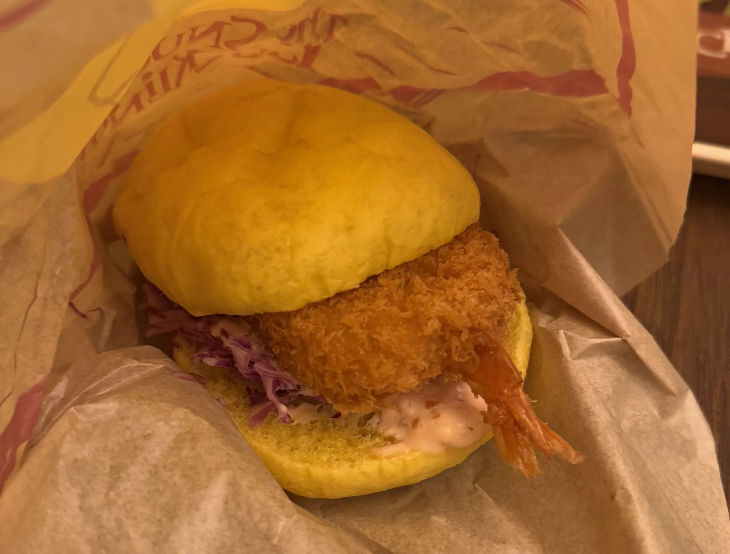 Fried shrimp patty with tail in between a yellow burger bun with shaved red cabbage and a pink orange sauce under the shrimp patty