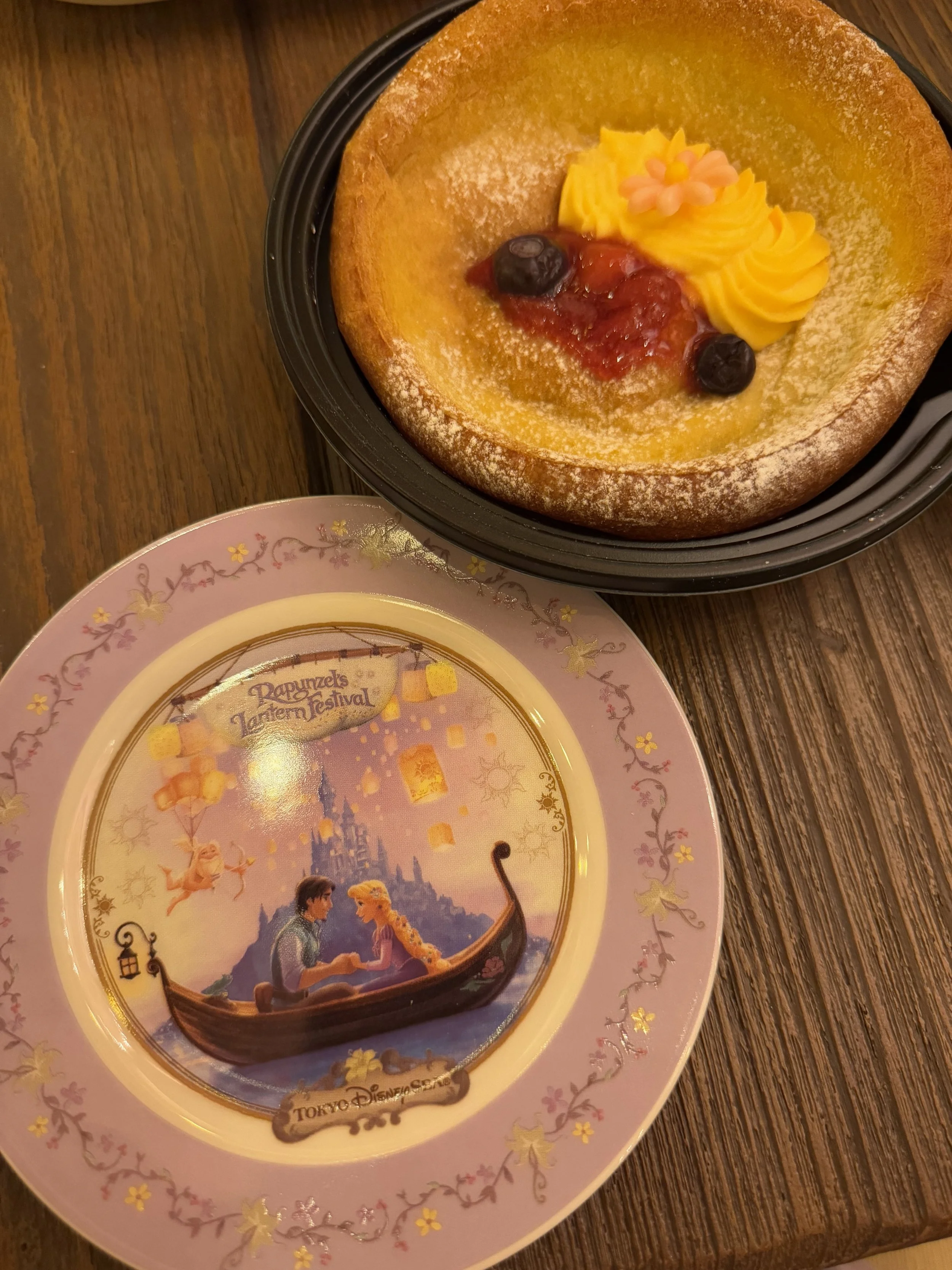 Pancake in black plastic tray with yellow cream and fruit jam next to a purple Rapunzel themed souvenir small plate