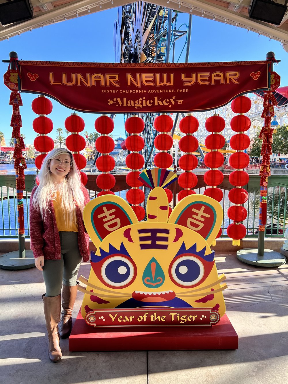  A girl with blond hair and a yellow top with red cardigan and green pants standing next to a cardboard image of a cartoon tiger with a Year of the Tiger banner at the bottom and a Lunar New Year banner at the top 