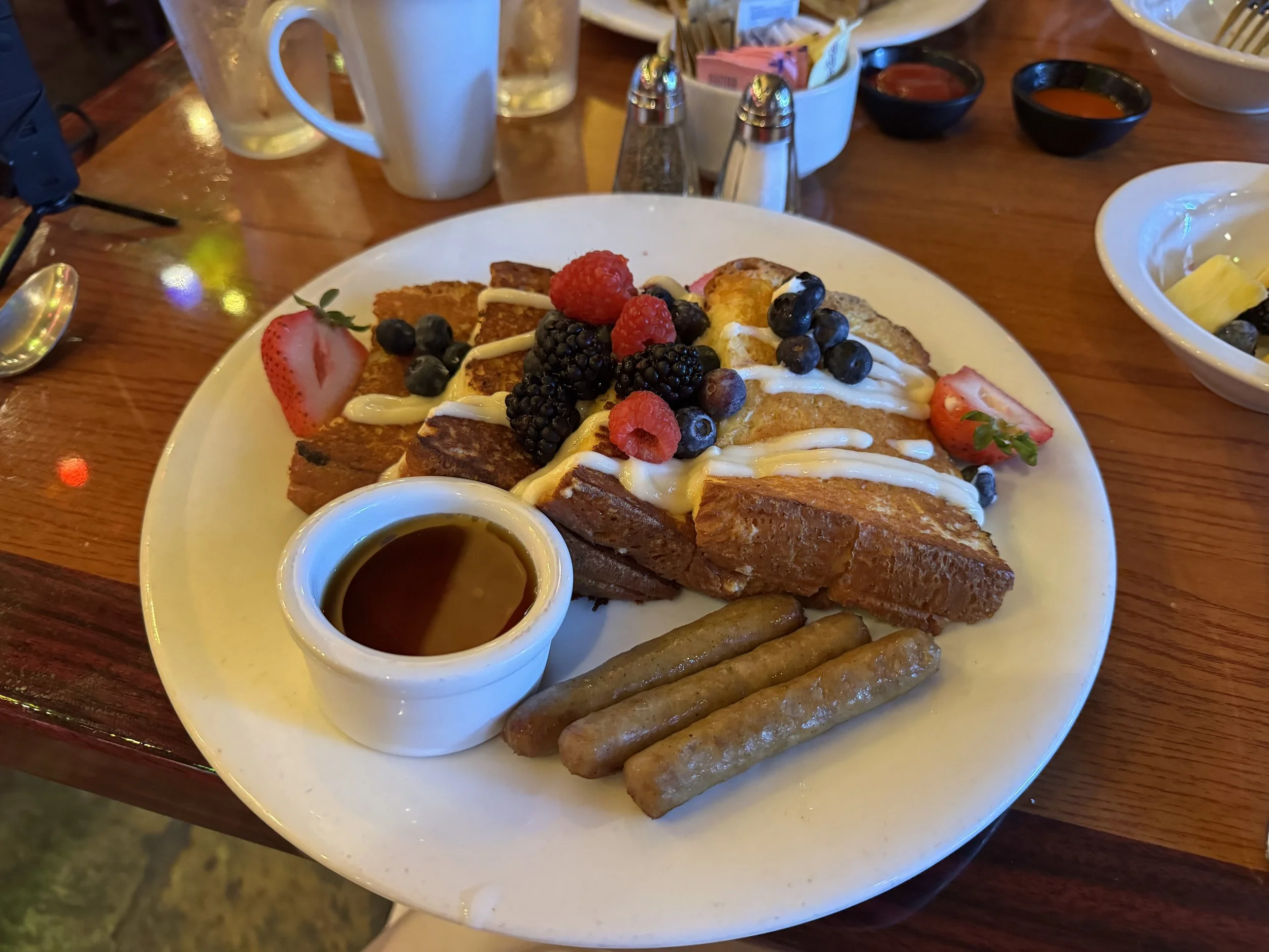  Plate with 4 large slices of French toast topped with fruits and cream cheese icing and 3 sausages with a small cup of syrup   
