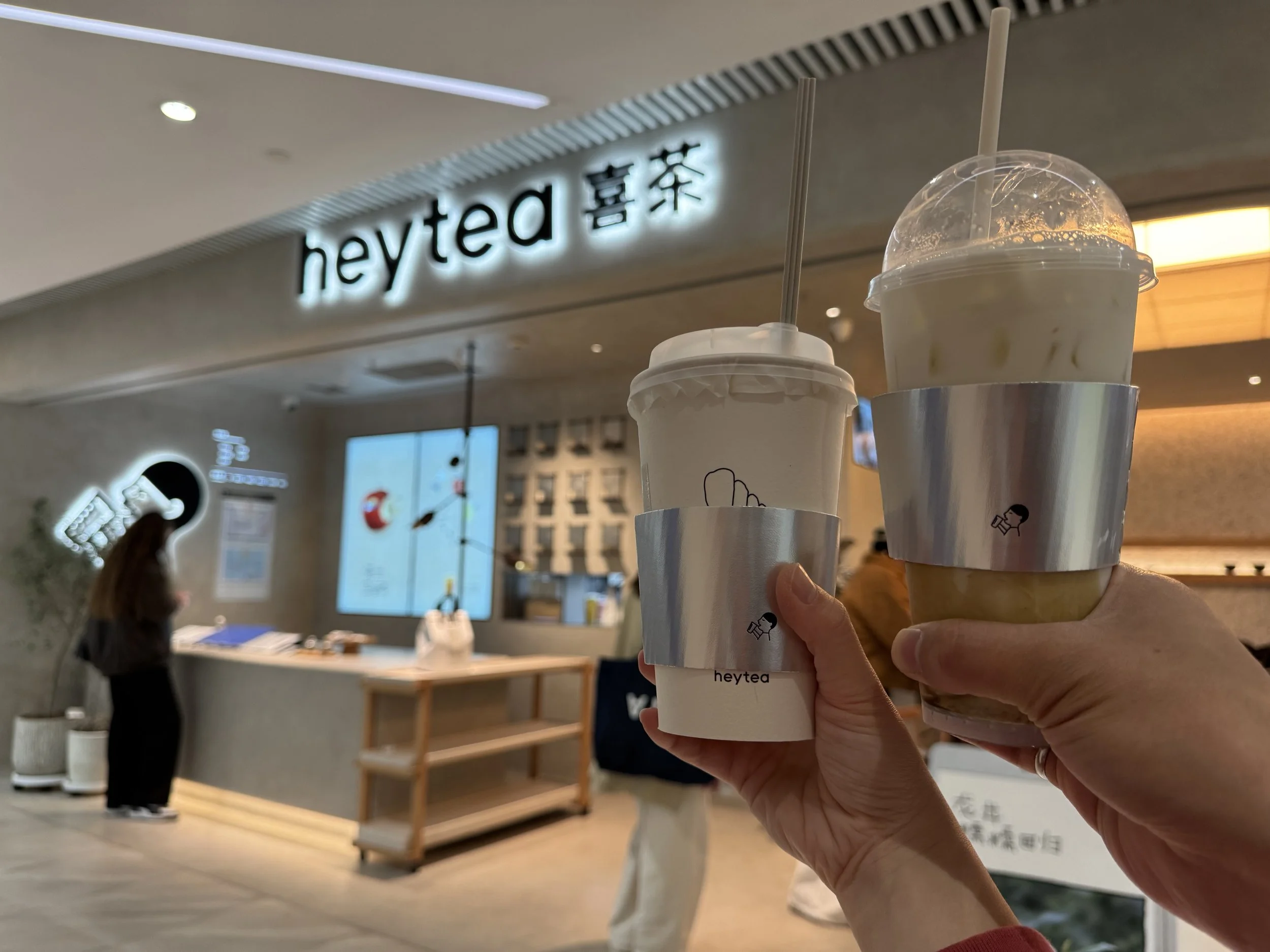 Two cups of milk tea in silver cup sleeves in front of a hey tea storefront