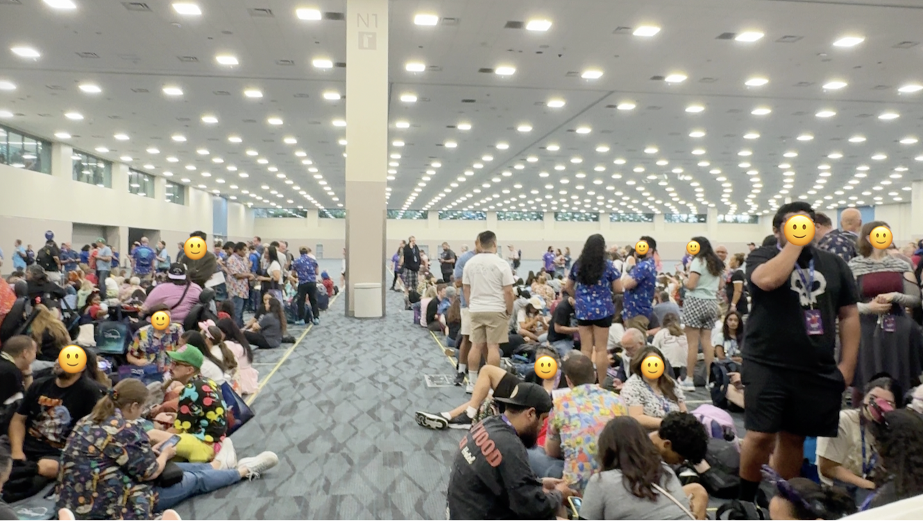 Rows of people sitting and standing in line inside a brightly lit large ballroom