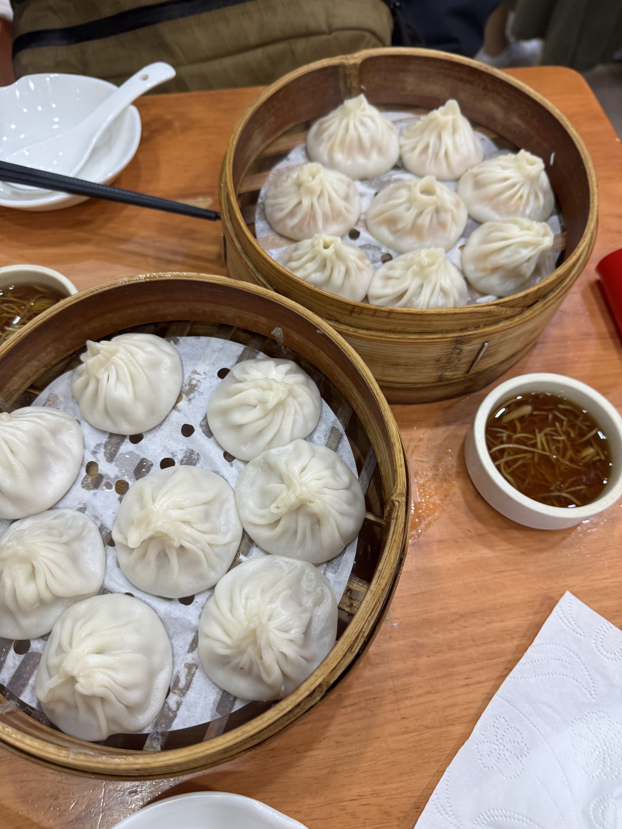 Crab and pork xialongbao in bamboo steamer trays on a wooden table with small saucers of vinegar and sliced ginger