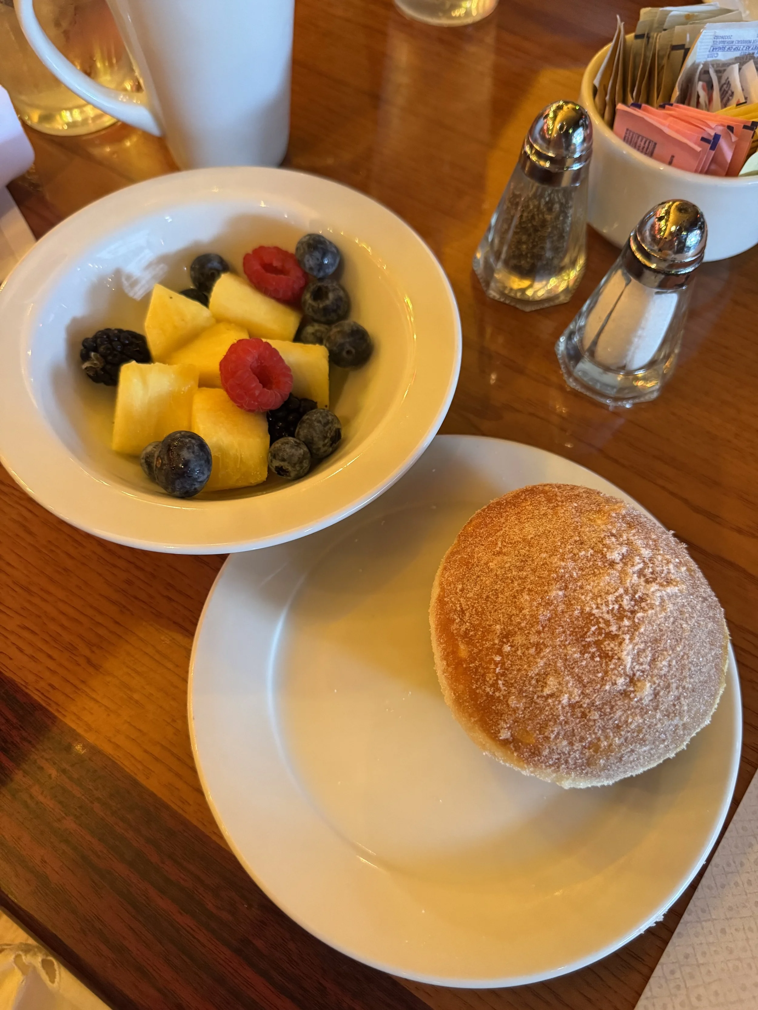 Small bowl of cut pineapple, blueberries, and raspberries next to a small plate of one large malasada