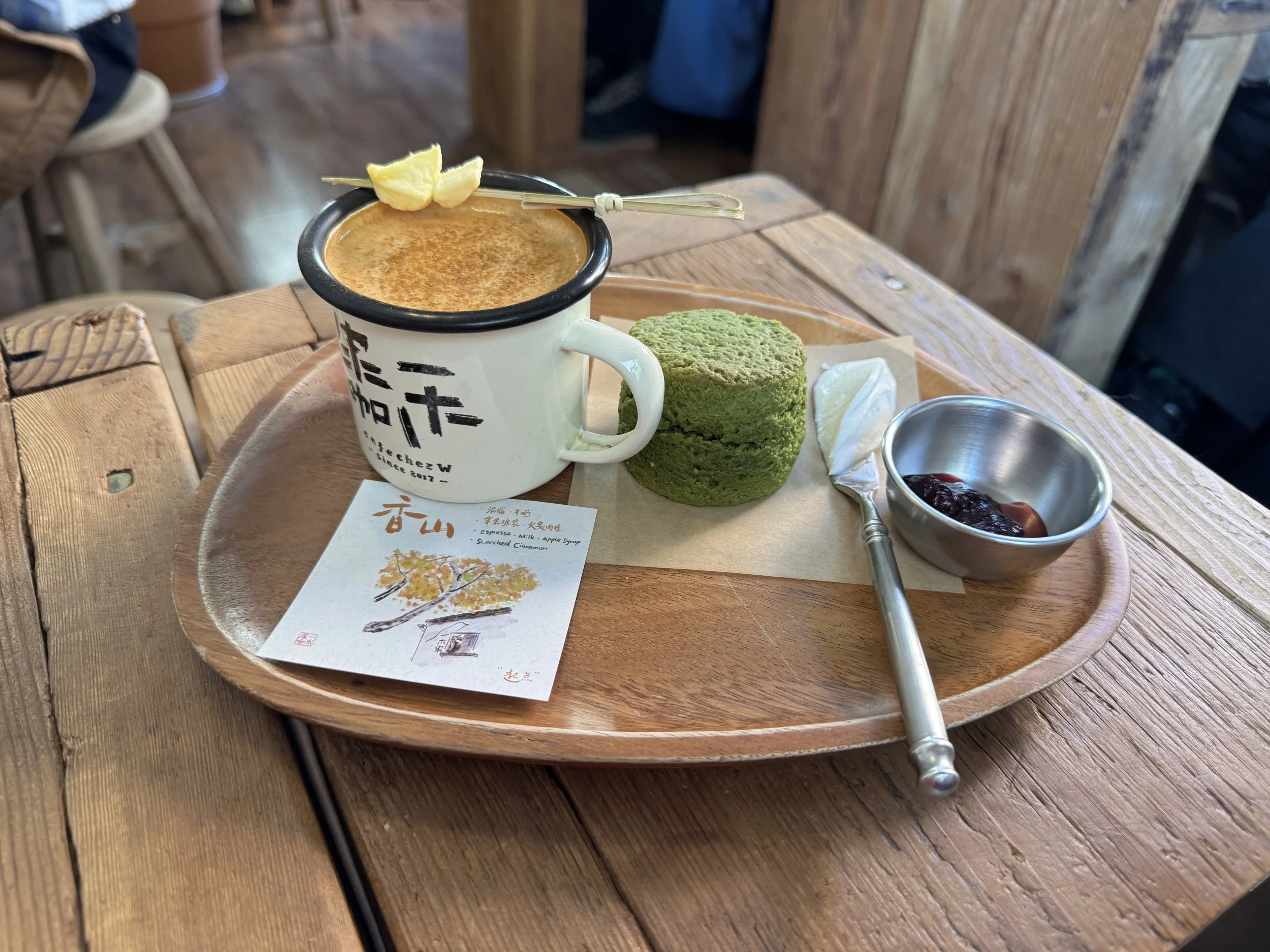 white mug of apple cinnamon latte with freeze dried apple pieces in a skewer on top next to a dark green matcha scone with jam and cream on a wooden plate