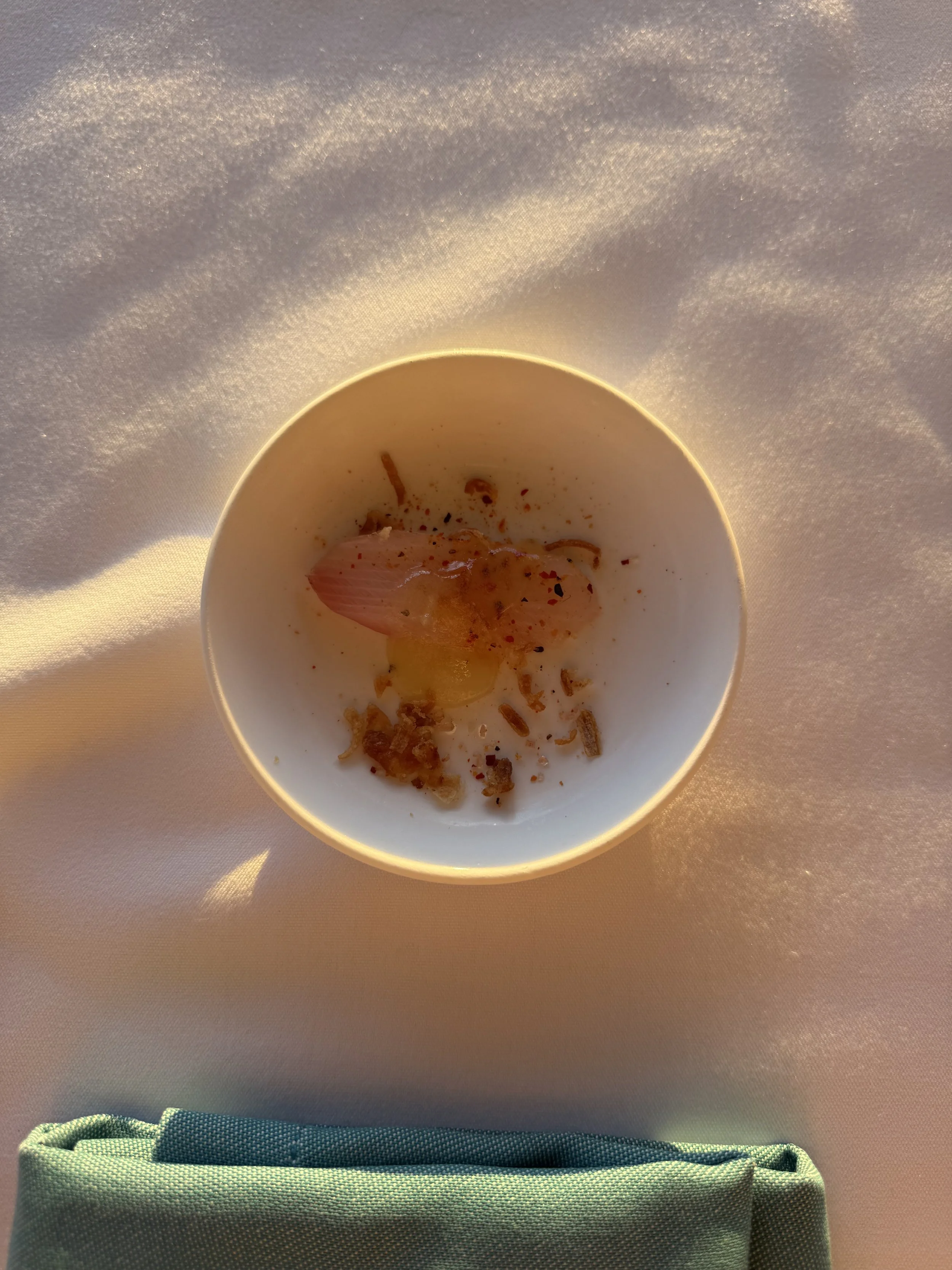 Top-down view of a slice of yellowtail sashimi with crispy onions on a small white circular plate on a table with white tablecloth