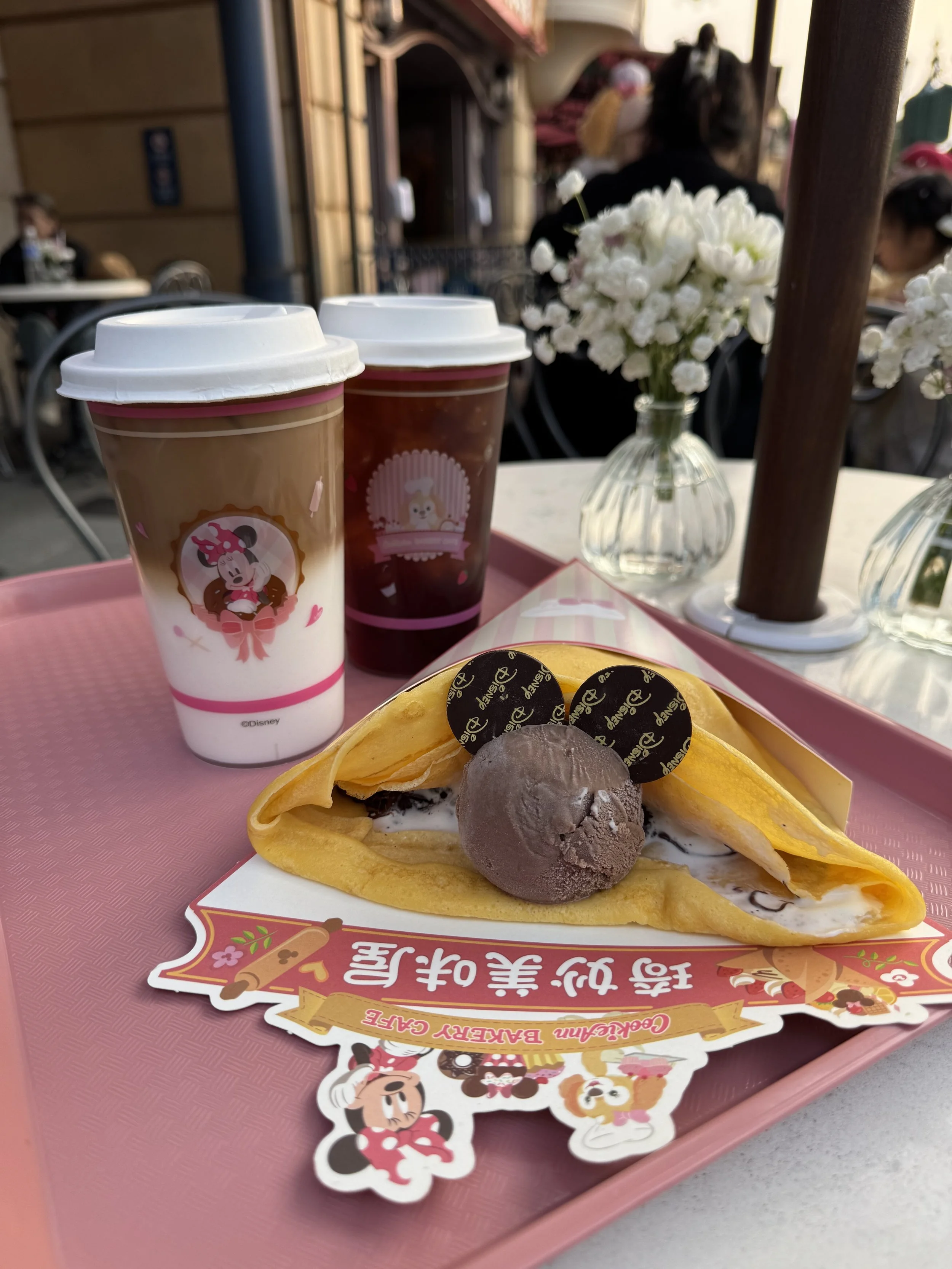 Crepe with chocolate ice cream and two Disney chocolate disks to look like Mickey Mouse next to two clear cups of coffee with Minnie Mouse and Cookie Ann on the sides
