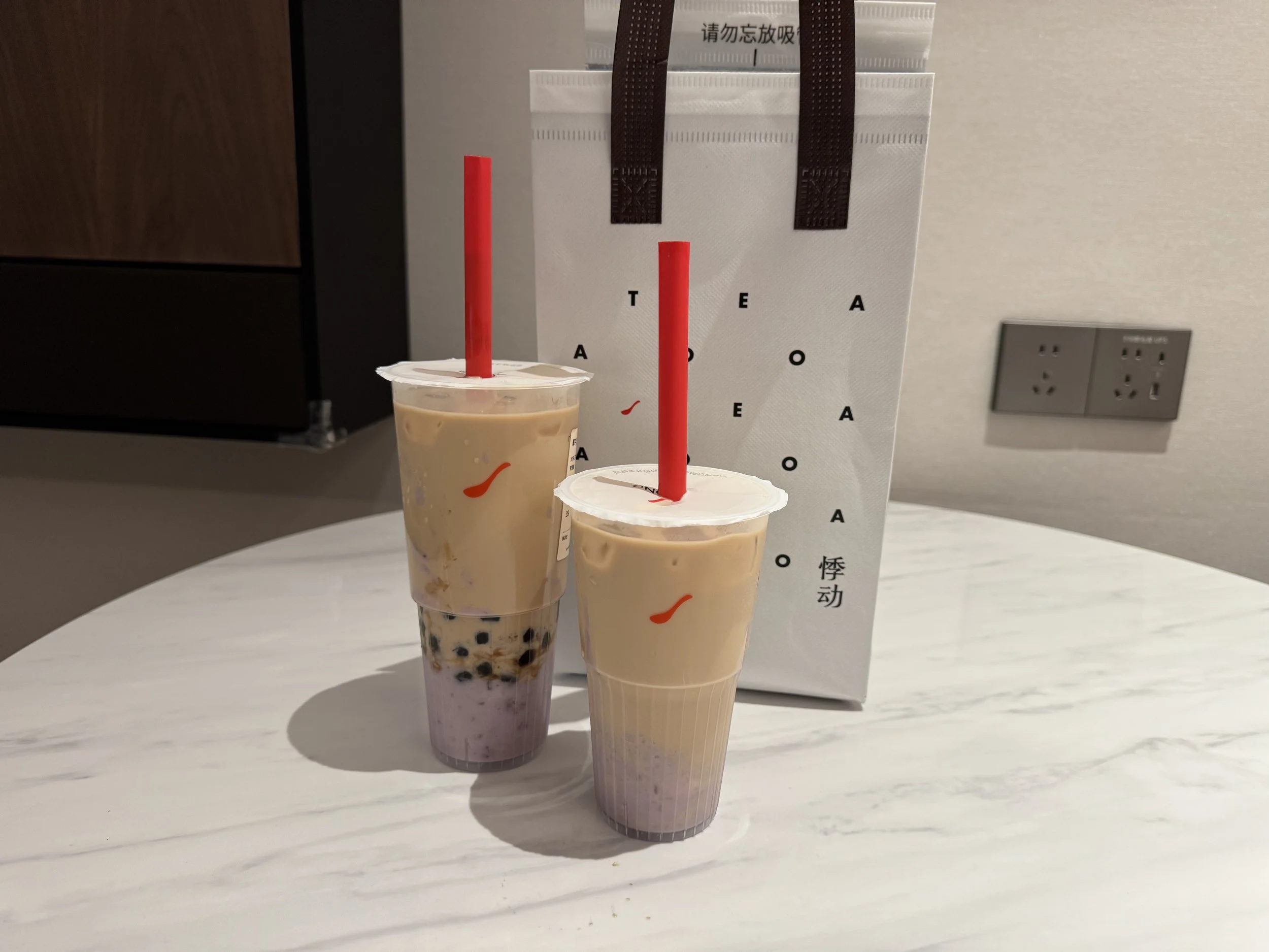 Two cups of milk tea with taro mash topping on a white table in front of an insulated bag