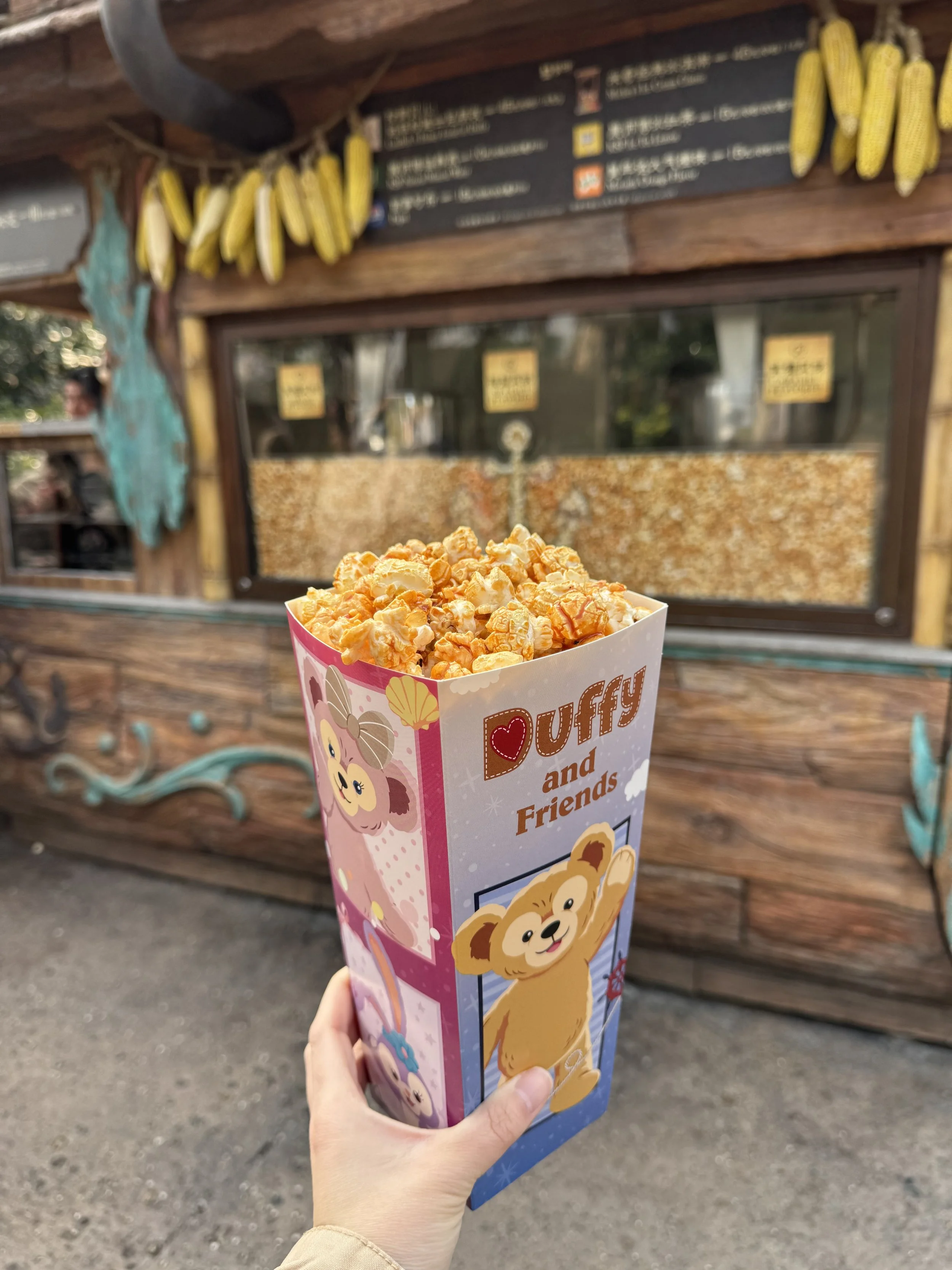Golden caramel popcorn in a Duffy and Friends cardboard popcorn box in front of a popcorn cart that has hanging corn from the roof and popcorn in a glass case