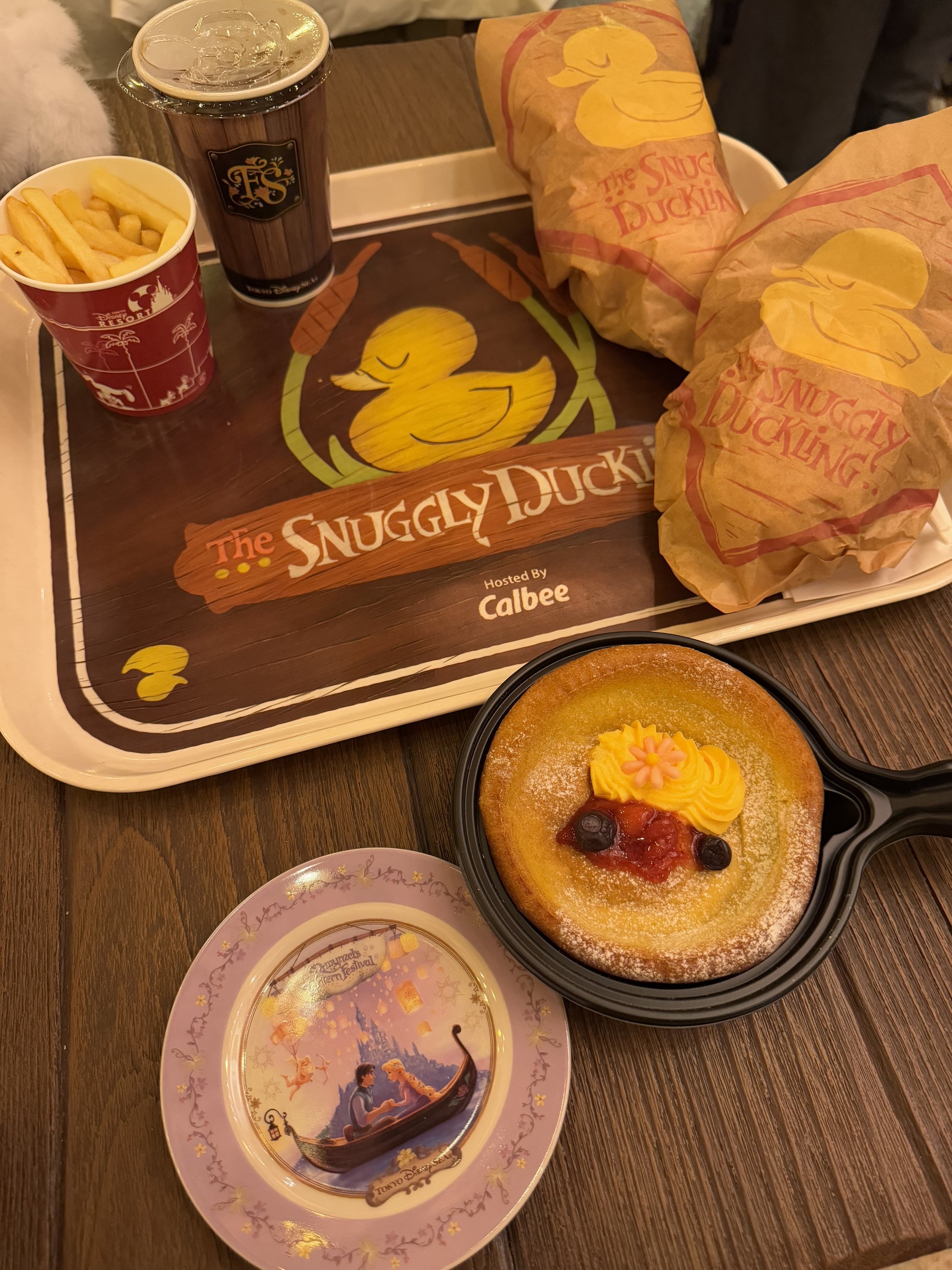 Picture of food spread on a plastic tray with a cup of fries, a brown cup, two burgers wrapped in parchment paper and a pancake on a black frying pan plastic tray and a purple small Rapunzel souvenir plate
