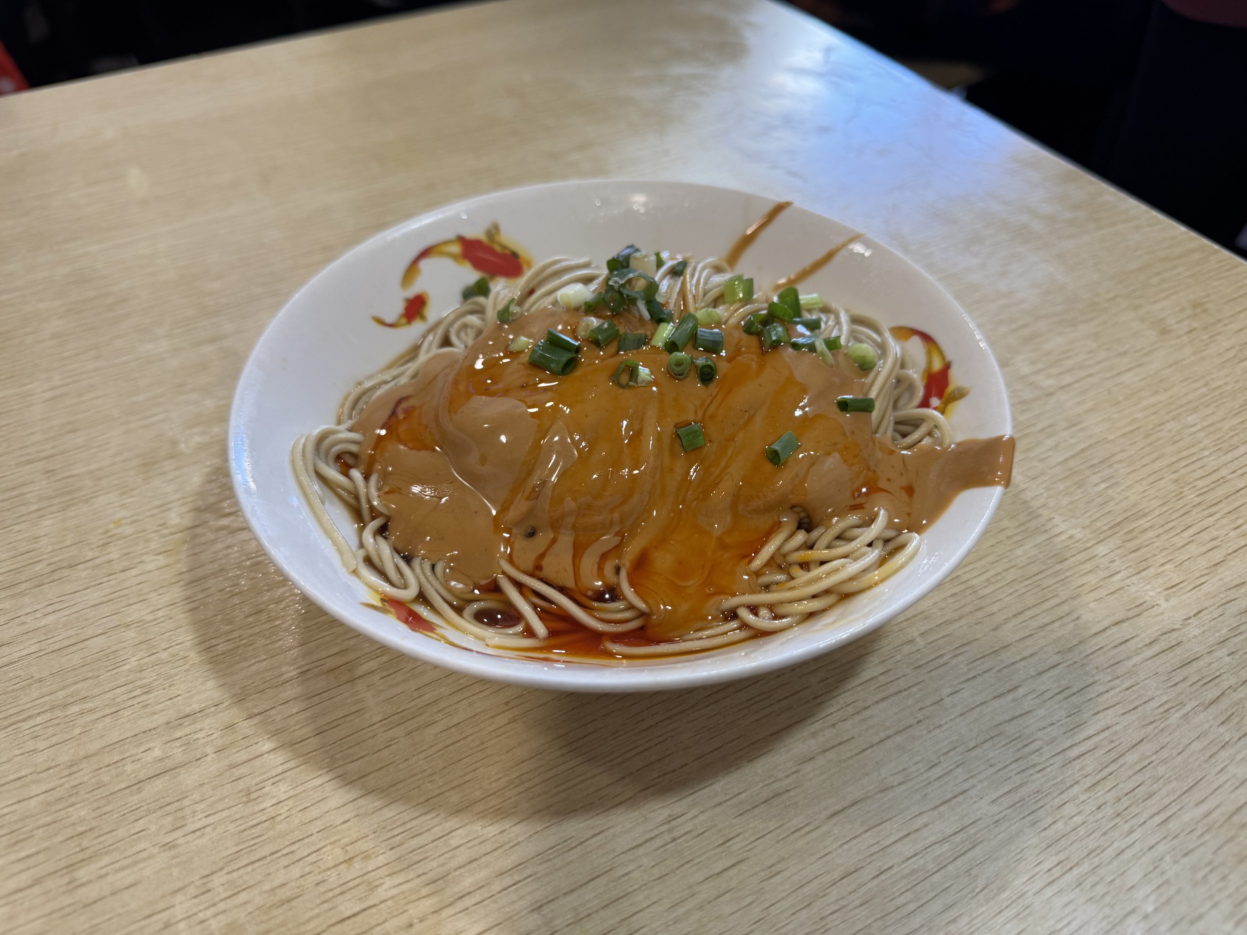 Small white bowl of noodles with brown sesame sauce and red chili oil and sliced green onions on top