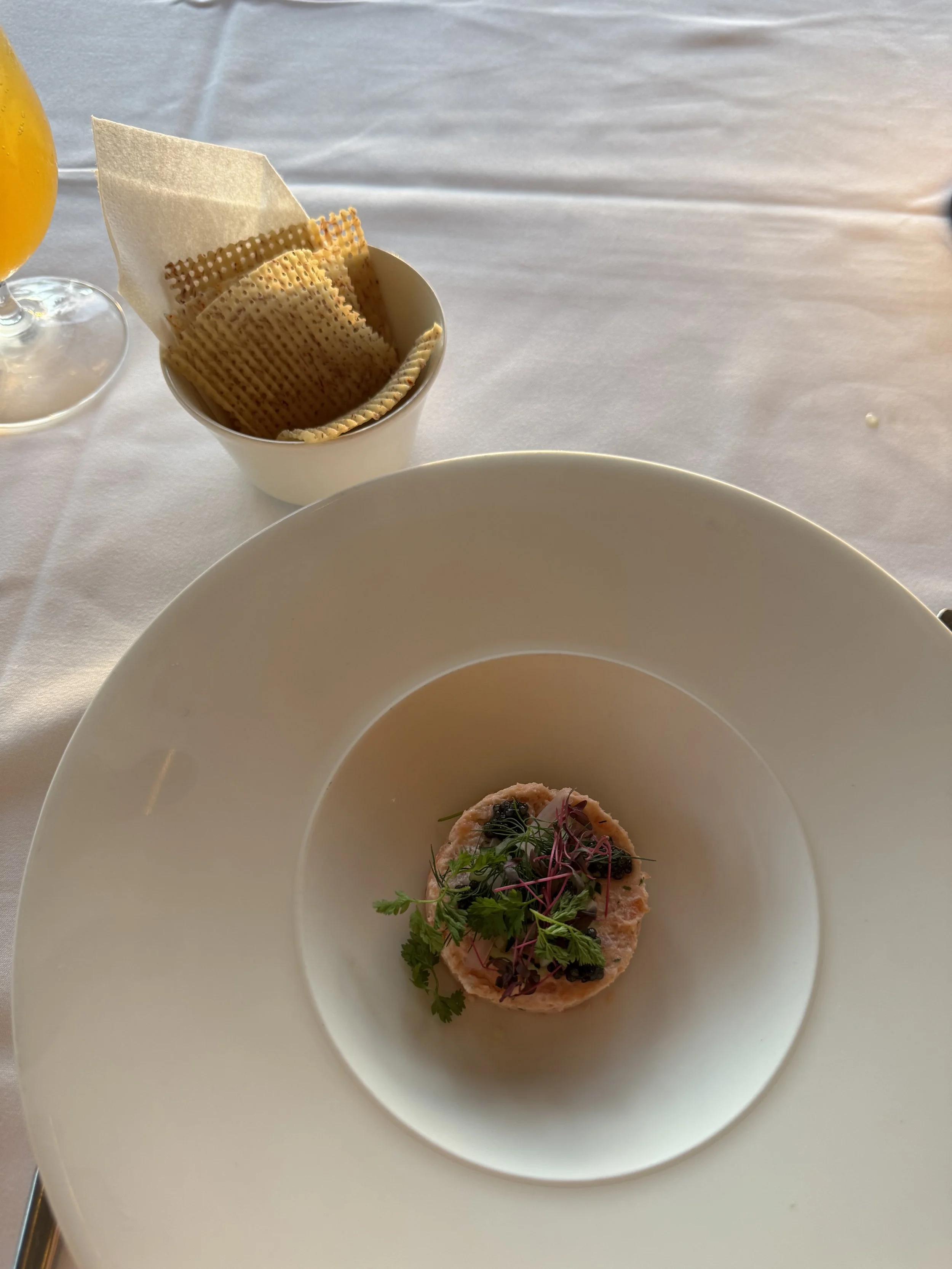 A cylindrical mound of pink smoked salmon pate with micro greens on top in a large white circular plate next to a white cup of weaved taro chips