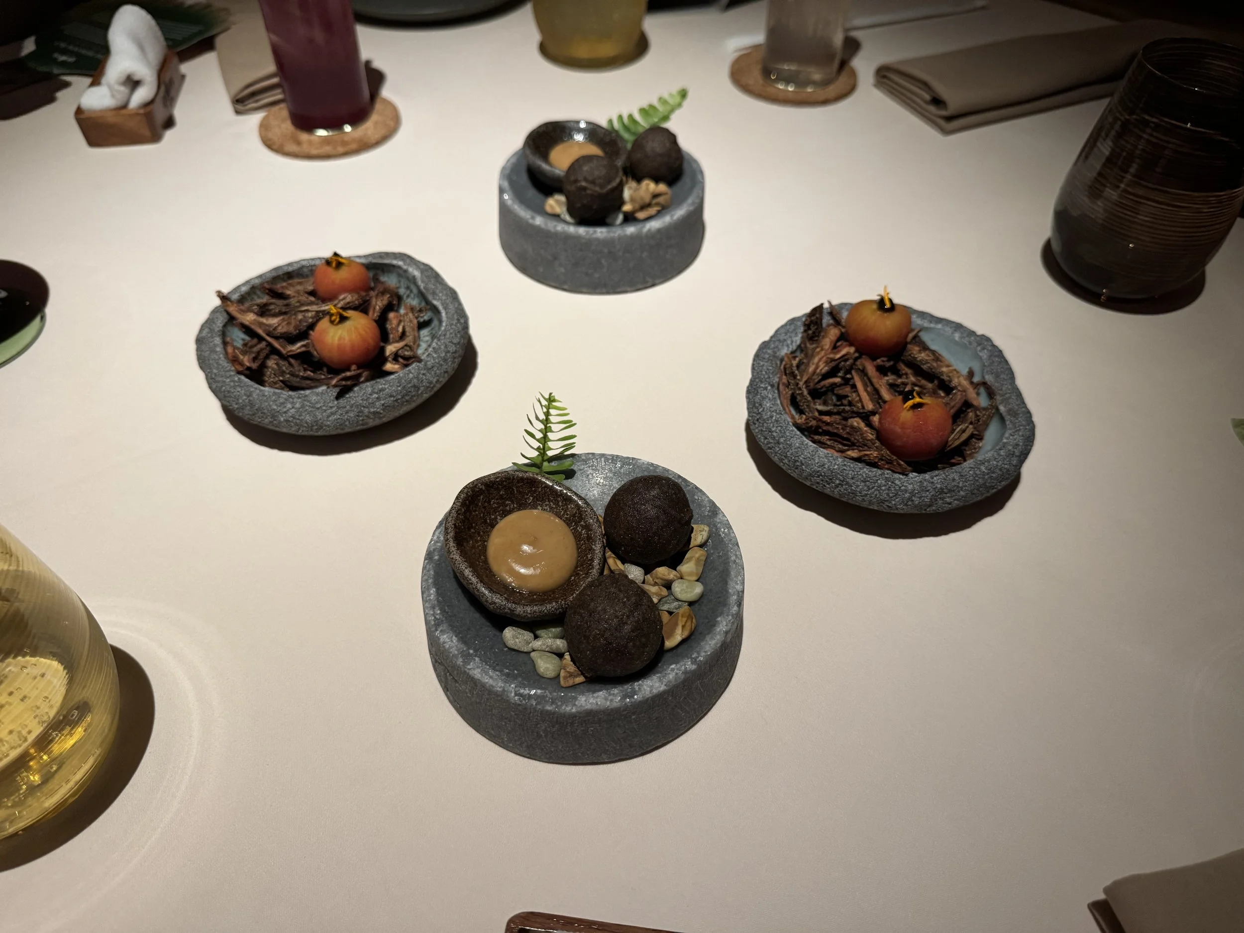 4 stone bowls with two tomatoes on a bed of bark and two black balls next to a small plate of brown sauce on a bed of small pebbles.