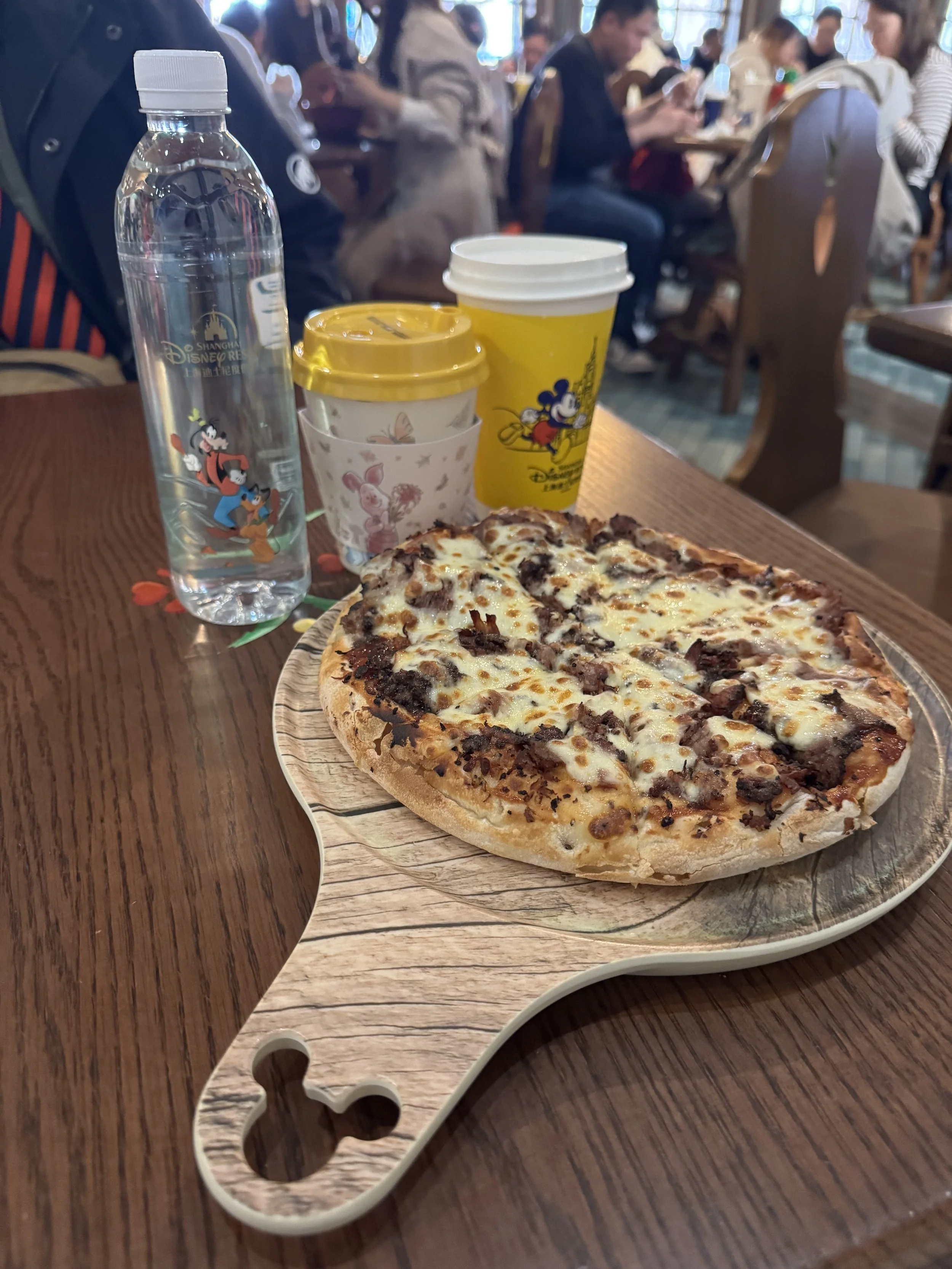 Pizza on a wooden platter with a Mickey shaped hole on the handle in front of a bottle of water, and two cups with Disney decorations on them