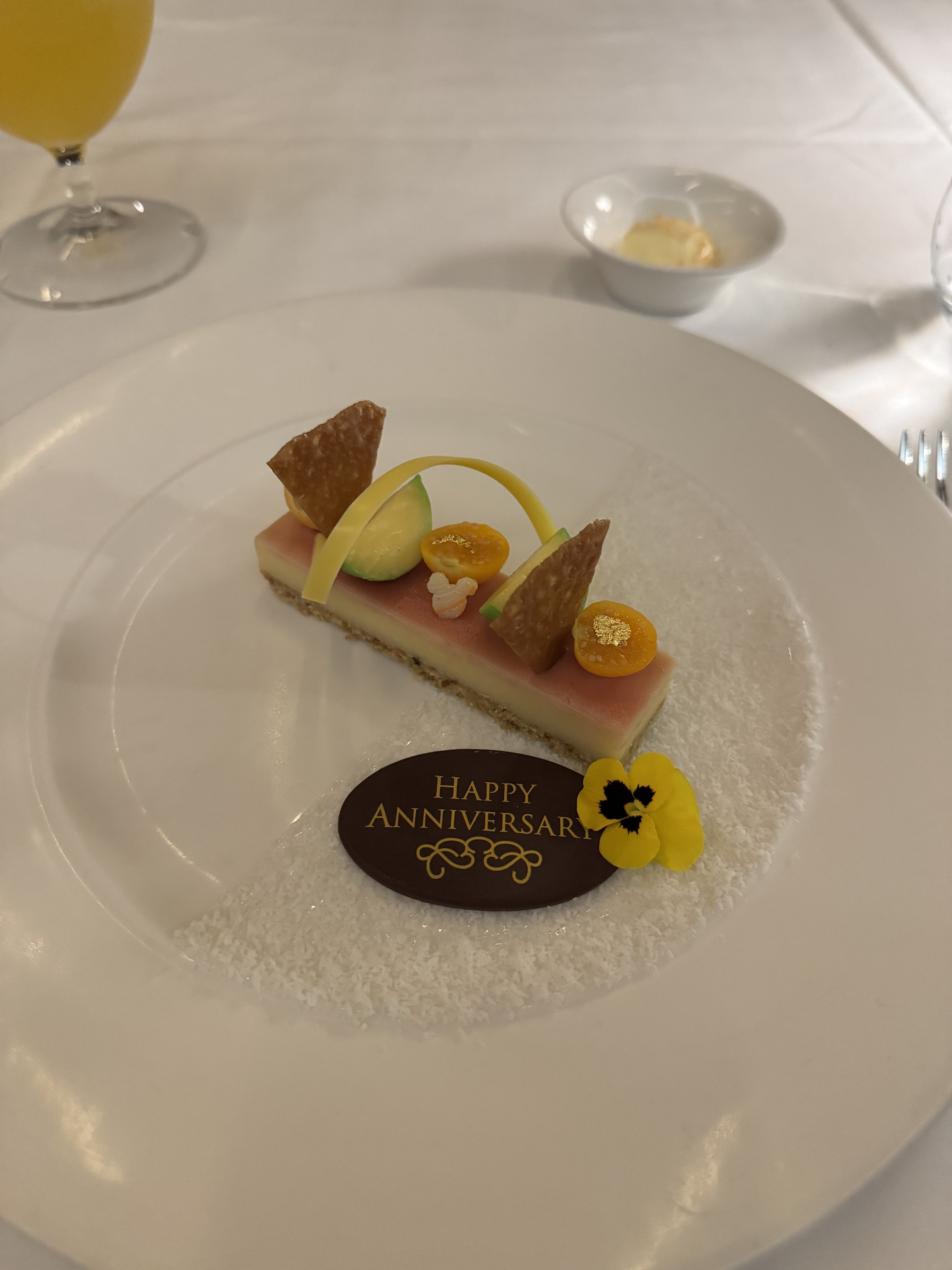 rectangular bar with a crunchy bottom layer, and a yellow and orange mousse layer topped with toffee slices cut into triangles, small oranges and calamansi mousse with a white chocolate curl on a white plate covered in shaved coconut and a chocolate