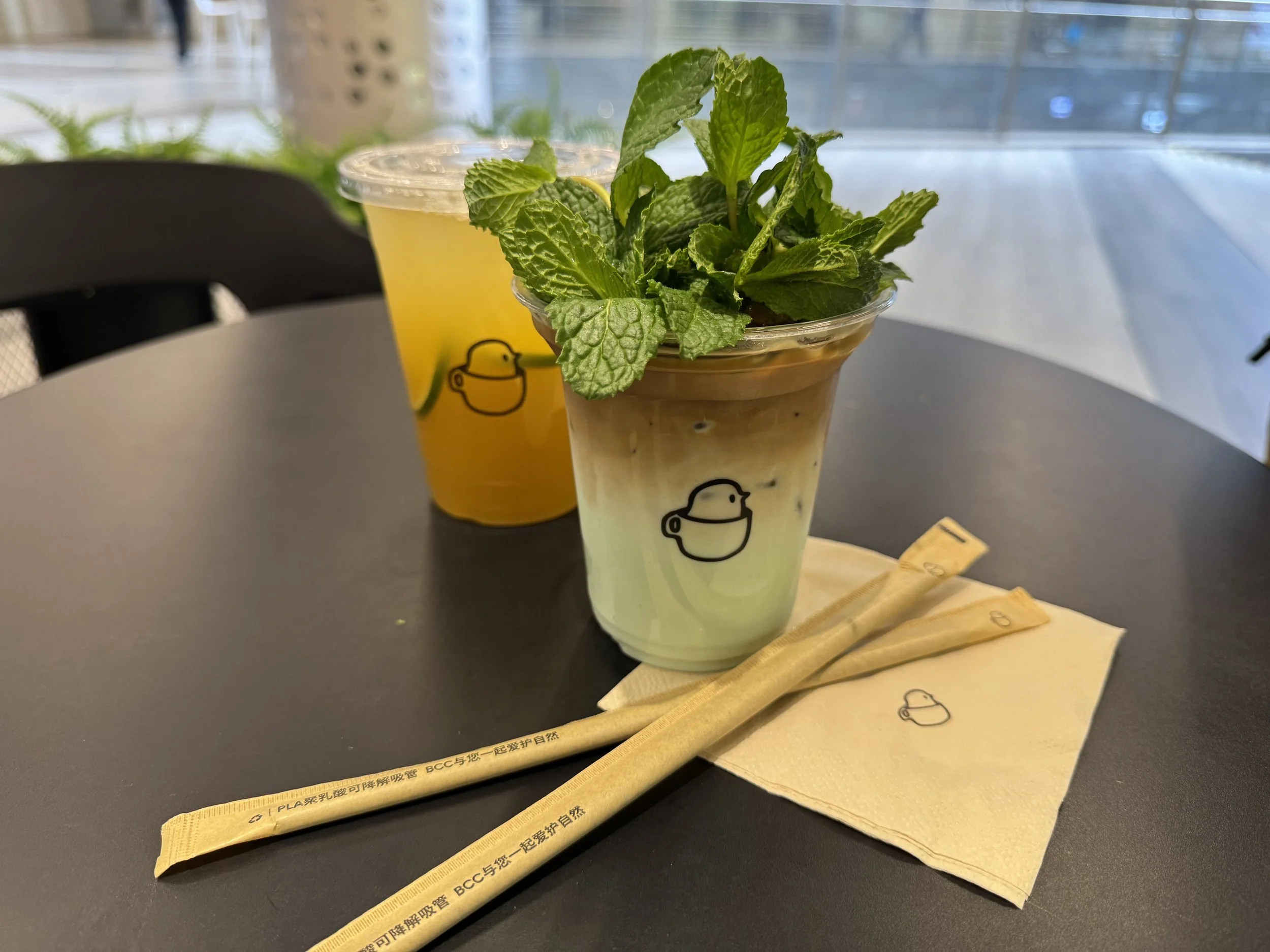 a cup of coffee on light green colored milk with a bundle of mint leaves on top in front of a cup of lemon jasmine tea on a black table with a square napkin and two straws