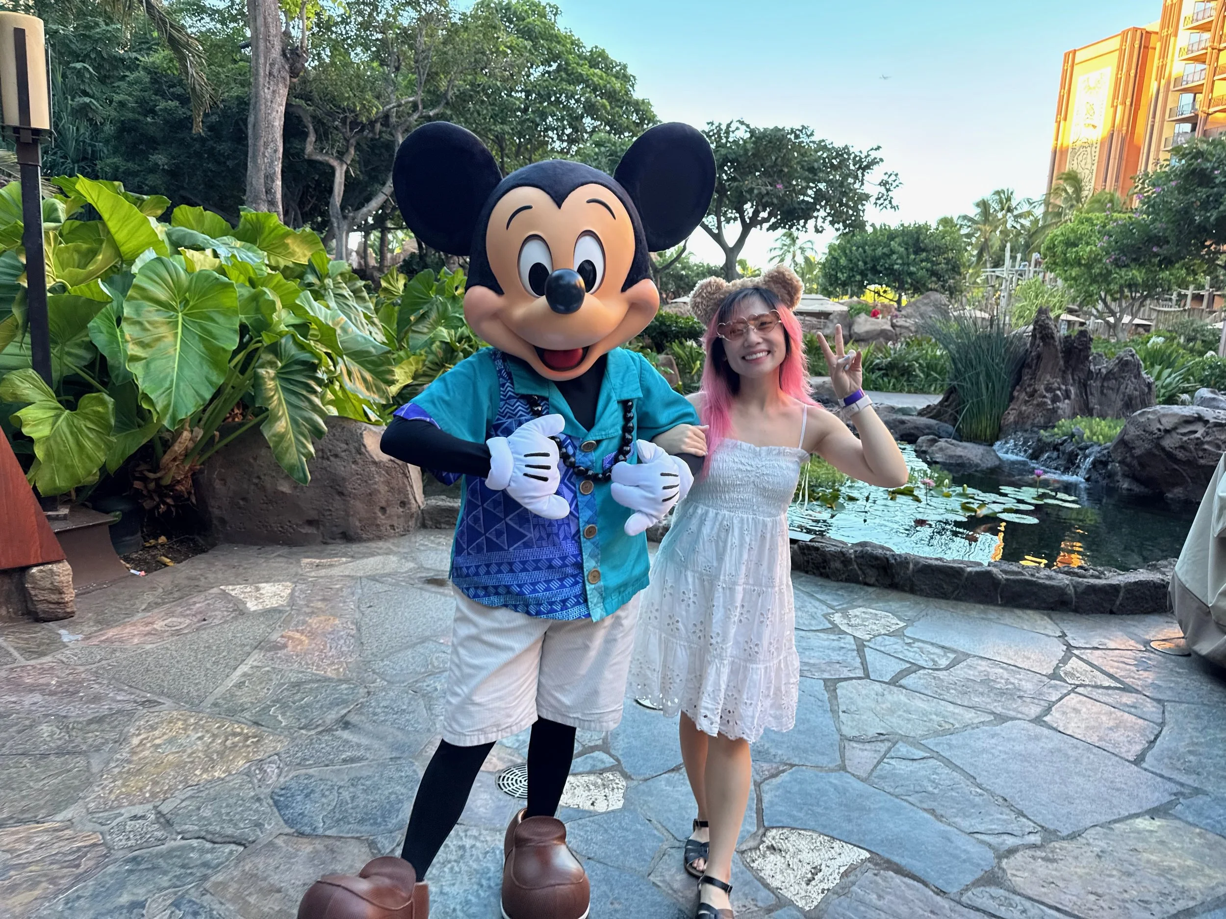 A girl with Duffy ears and Duffy sunglasses linking arms with Mickey Mouse in a Hawaiian shirt and white shorts at Aulani