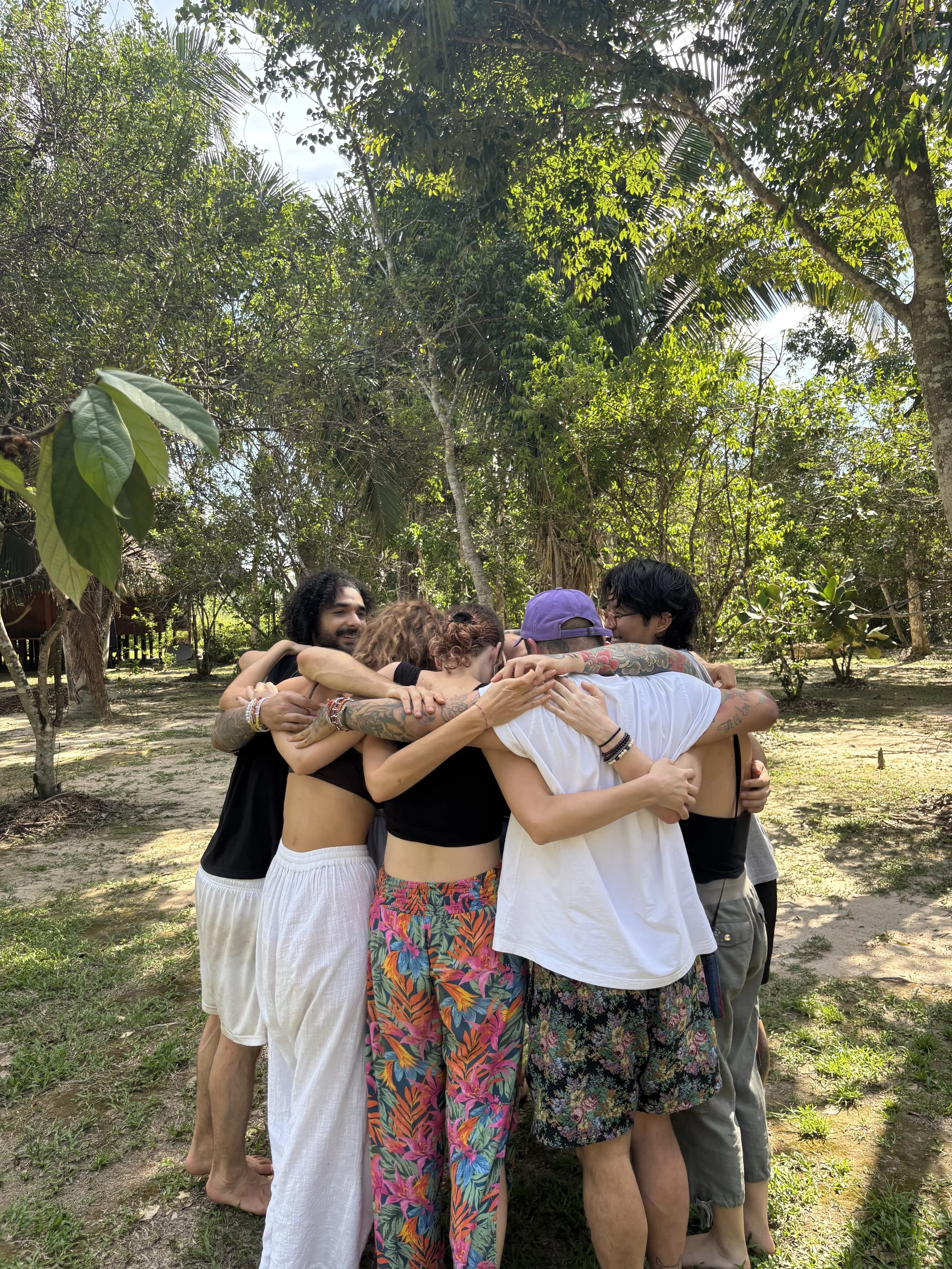 Group of five people hugging outdoors in a lush, tropical forest setting during daytime.