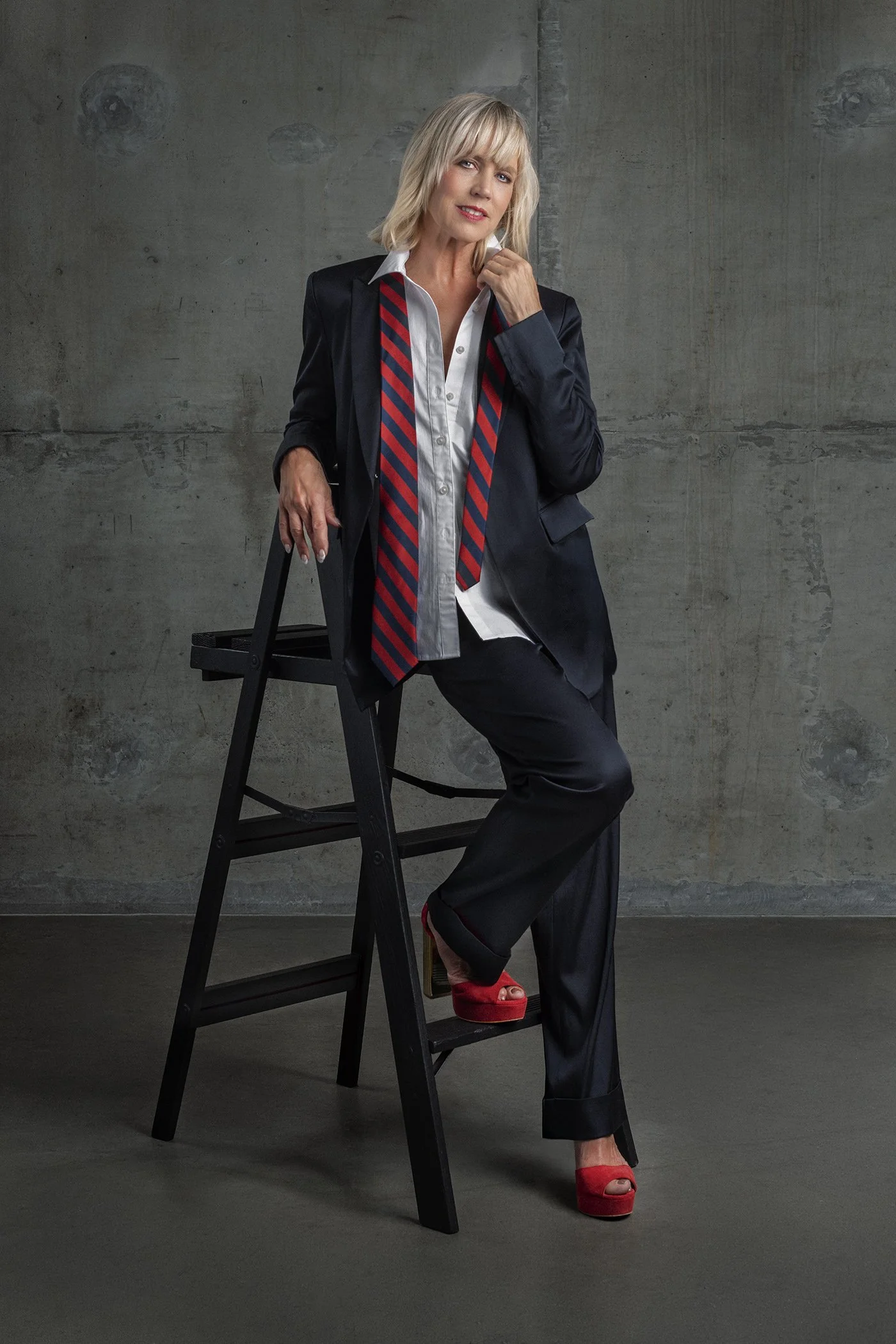 Catherine Grace O'Connell with blonde hair photographed in studio wearing navy blue suit and blazer and red and blue striped men's tie. Suit by Elaine Kim photographed in Seattle, Los Angeles and New York. Ageless in Seattle
