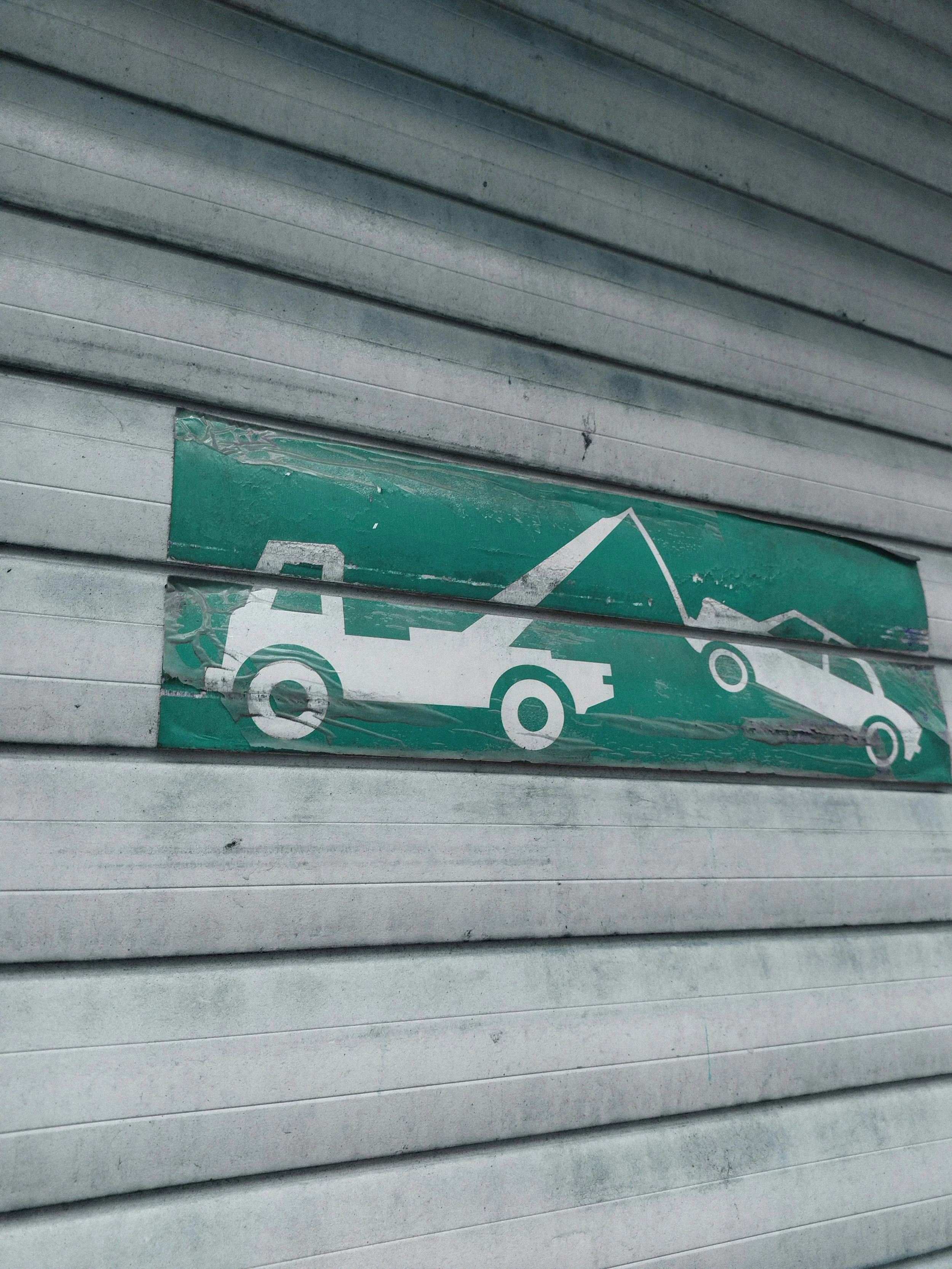Image of a scratched and worn sign showing a tow truck hooking a car, with mountains in the background, mounted on a metal rolling shutter door.